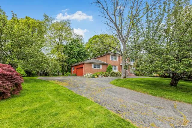 Brick exterior residence with a red garage door, featuring a curved driveway, expansive green lawn, mature trees, and established shrubbery Brick exterior residence with a red garage door, featuring a curved driveway, expansive green lawn, mature trees, and established shrubbery
