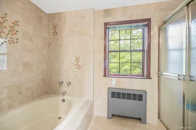 Tiled bathroom featuring a built-in bathtub with polished chrome fixtures, a window with a dark wood frame, a glass-enclosed shower, and a floor-mounted heating unit Tiled bathroom featuring a built-in bathtub with polished chrome fixtures, a window with a dark wood frame, a glass-enclosed shower, and a floor-mounted heating unit