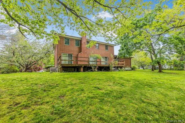 Expansive grassy backyard featuring a two-story brick exterior, a large wooden deck, multiple sliding glass doors, and mature trees providing natural shade Expansive grassy backyard featuring a two-story brick exterior, a large wooden deck, multiple sliding glass doors, and mature trees providing natural shade