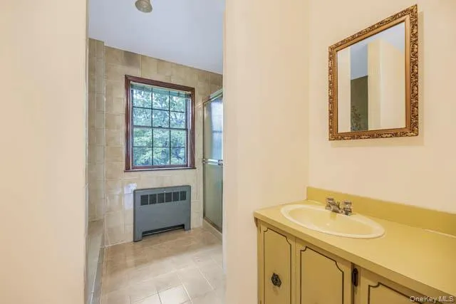 Bathroom featuring a vanity with an integrated sink, a framed wall mirror, a tiled shower enclosure, a window, and a baseboard heating unit Bathroom featuring a vanity with an integrated sink, a framed wall mirror, a tiled shower enclosure, a window, and a baseboard heating unit
