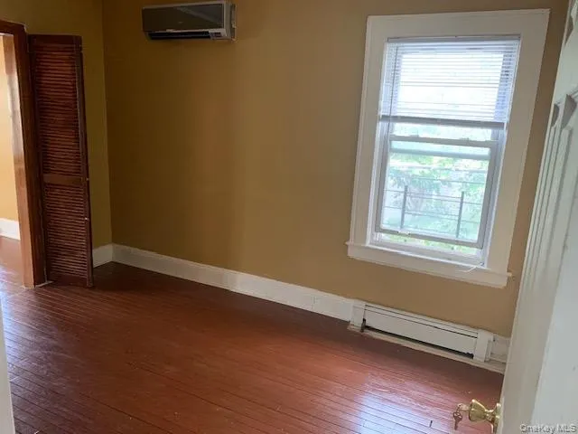 Empty room with baseboard heating, a wall mounted air conditioner, and wood-type flooring Empty room with baseboard heating, a wall mounted air conditioner, and wood-type flooring