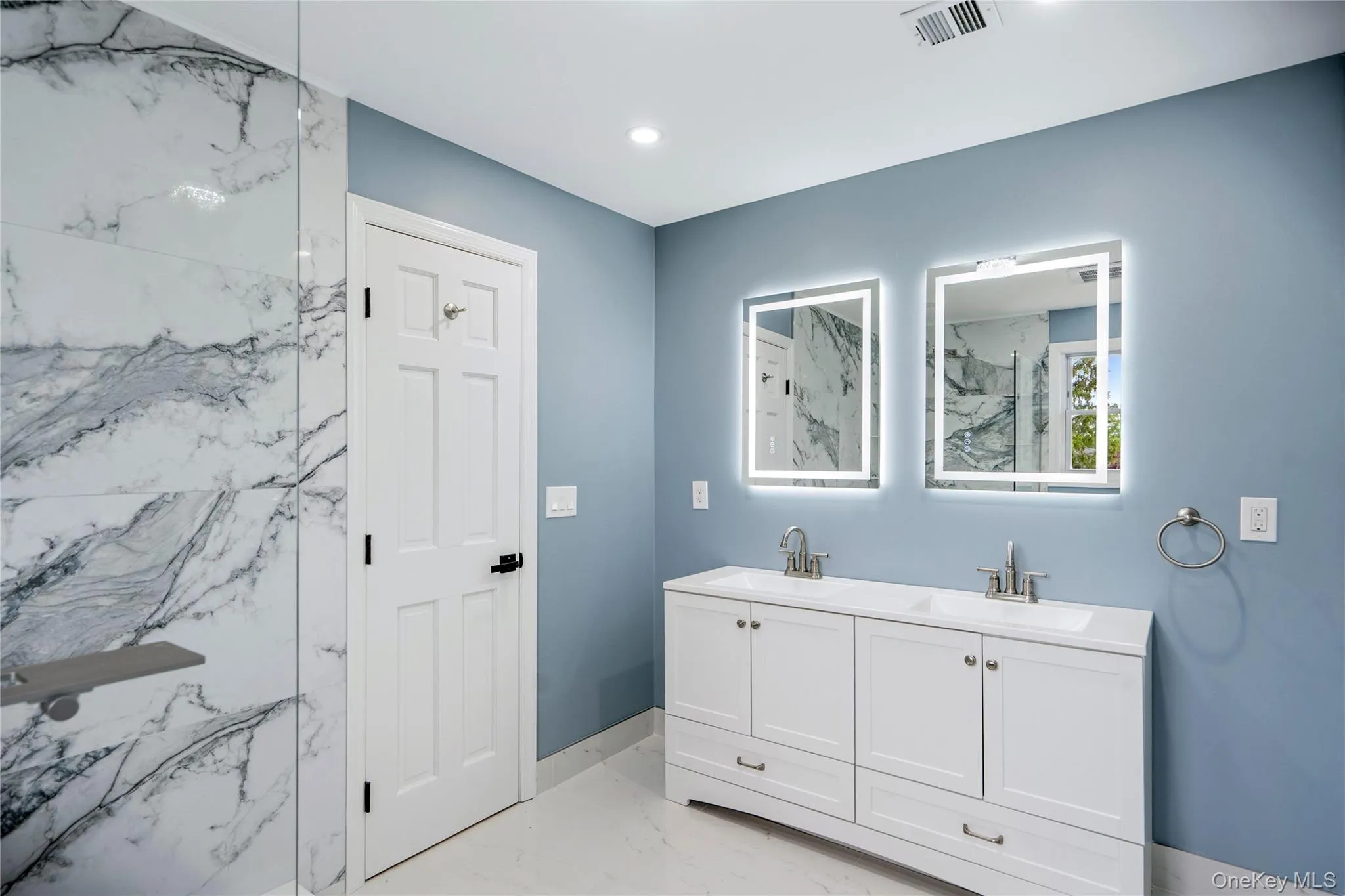 Modern en-suite bathroom featuring a double vanity with white cabinetry and integrated sinks, illuminated by two LED mirrors Modern en-suite bathroom featuring a double vanity with white cabinetry and integrated sinks, illuminated by two LED mirrors