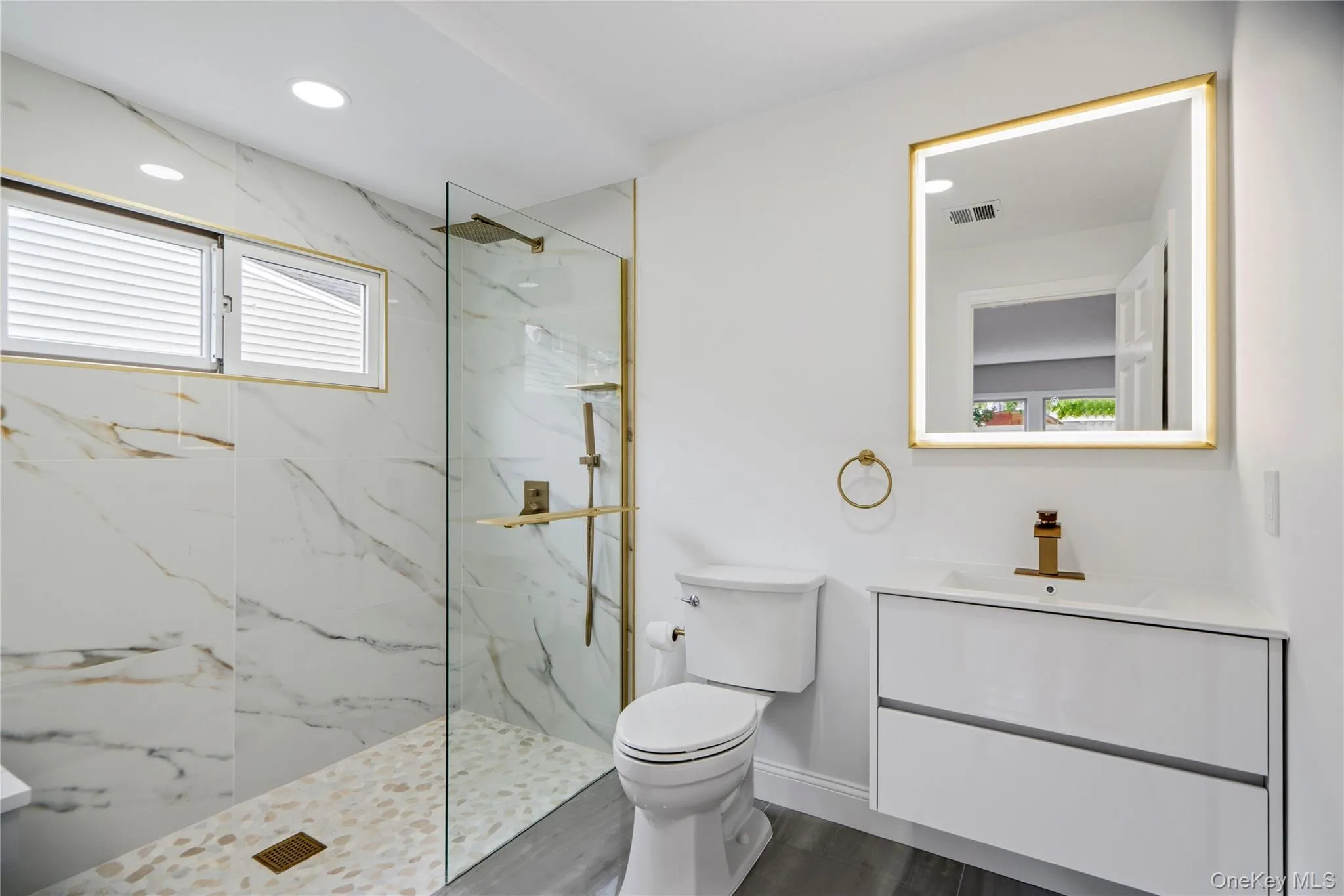Bathroom en-suite featuring a frameless glass shower enclosure with gold-finish hardware, marble-look tiled walls, pebble shower pan, and a floating vanity with an integrated sink Bathroom en-suite featuring a frameless glass shower enclosure with gold-finish hardware, marble-look tiled walls, pebble shower pan, and a floating vanity with an integrated sink