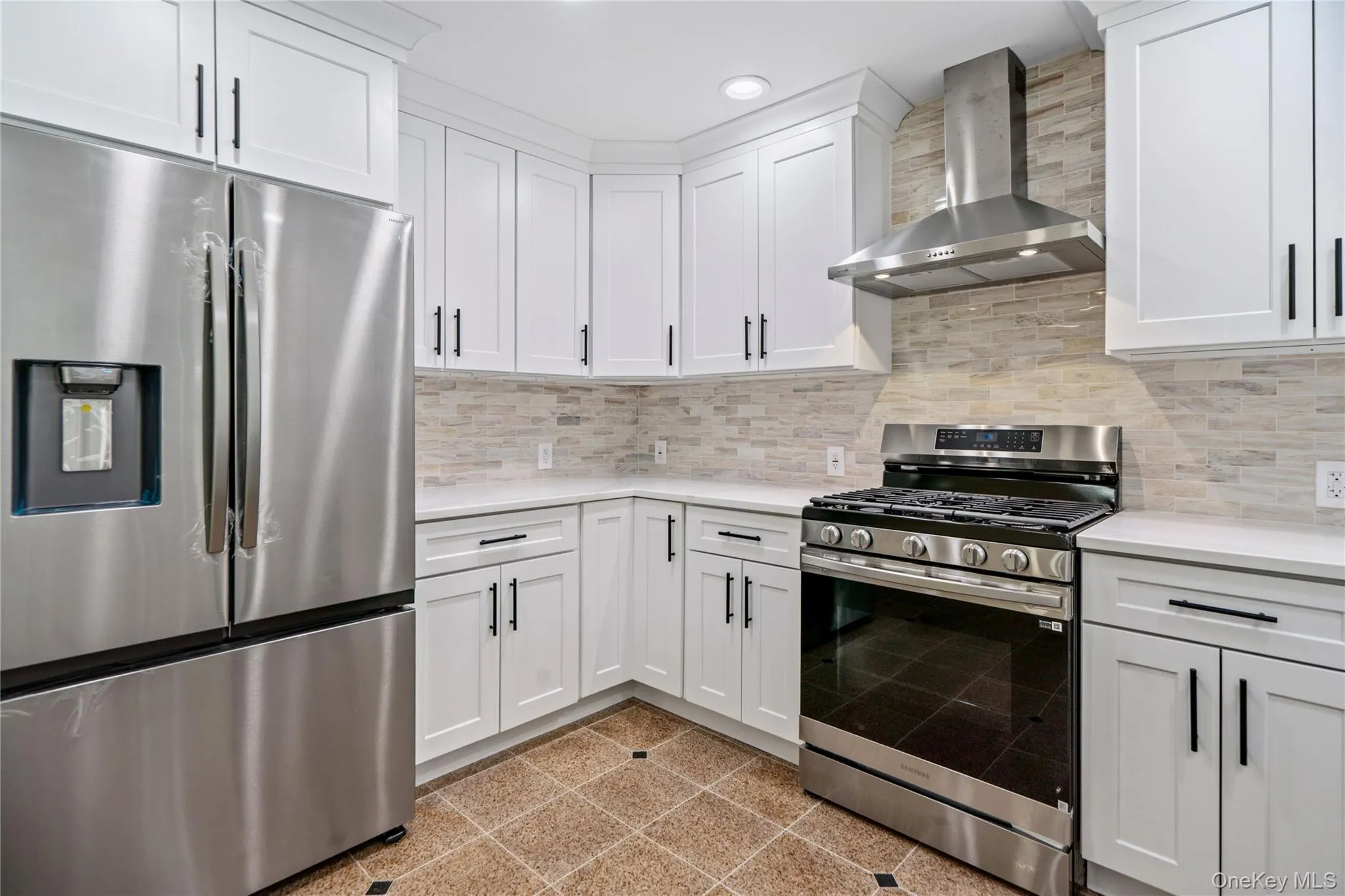 Modern kitchen featuring white shaker cabinetry with black hardware, stainless steel appliances, light-toned stone backsplash, and light-toned countertops Modern kitchen featuring white shaker cabinetry with black hardware, stainless steel appliances, light-toned stone backsplash, and light-toned countertops