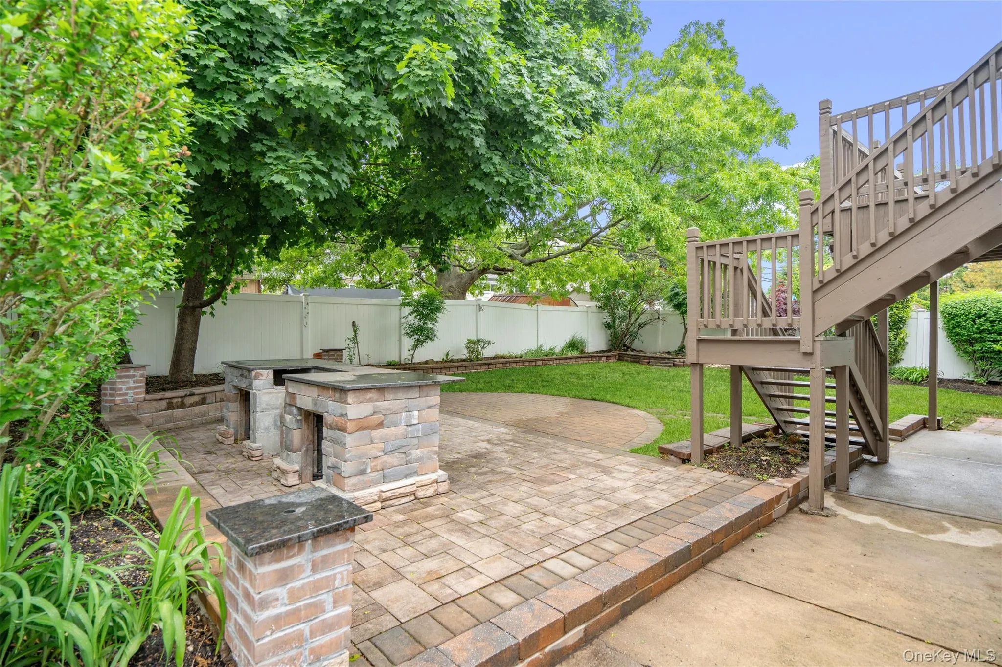 Paved patio area featuring two stone-clad built-in structures, a tiered wooden deck with railings and staircase, and a manicured lawn Paved patio area featuring two stone-clad built-in structures, a tiered wooden deck with railings and staircase, and a manicured lawn
