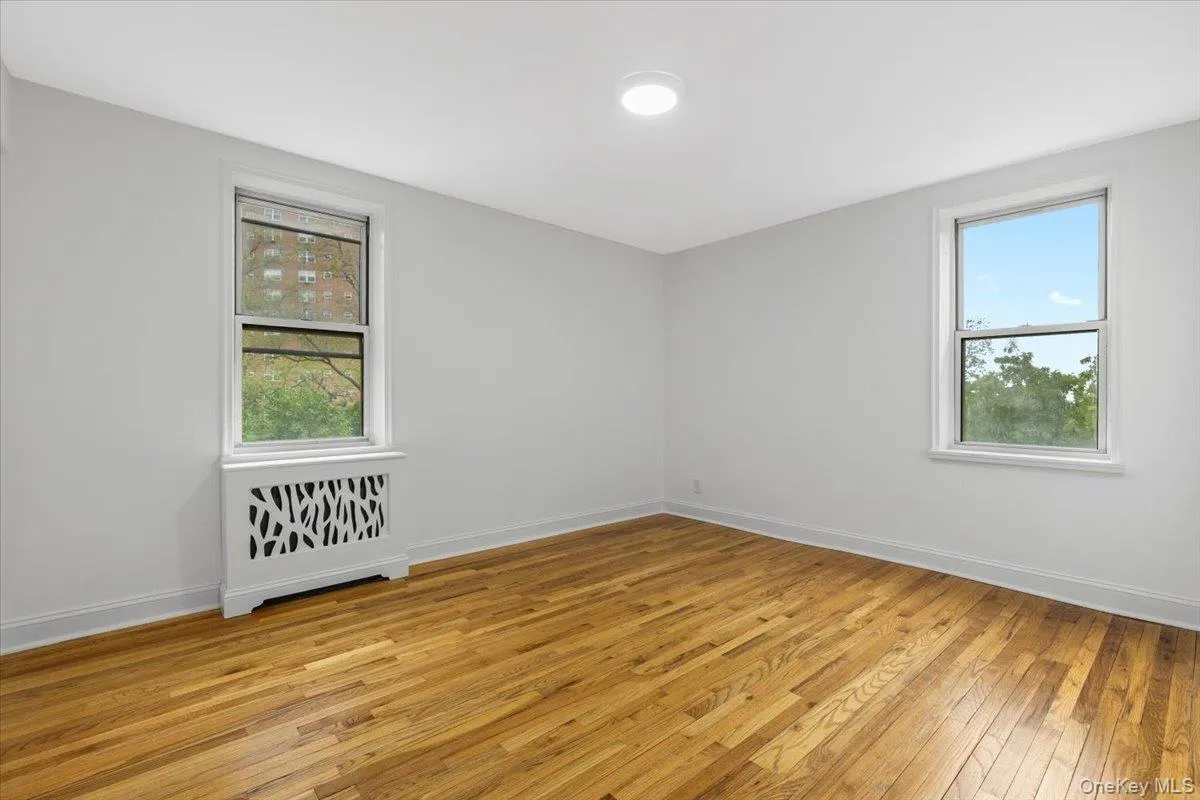 Hardwood flooring with a natural finish, two large windows, white radiator cover with decorative cutouts, and a flush-mount ceiling light Hardwood flooring with a natural finish, two large windows, white radiator cover with decorative cutouts, and a flush-mount ceiling light