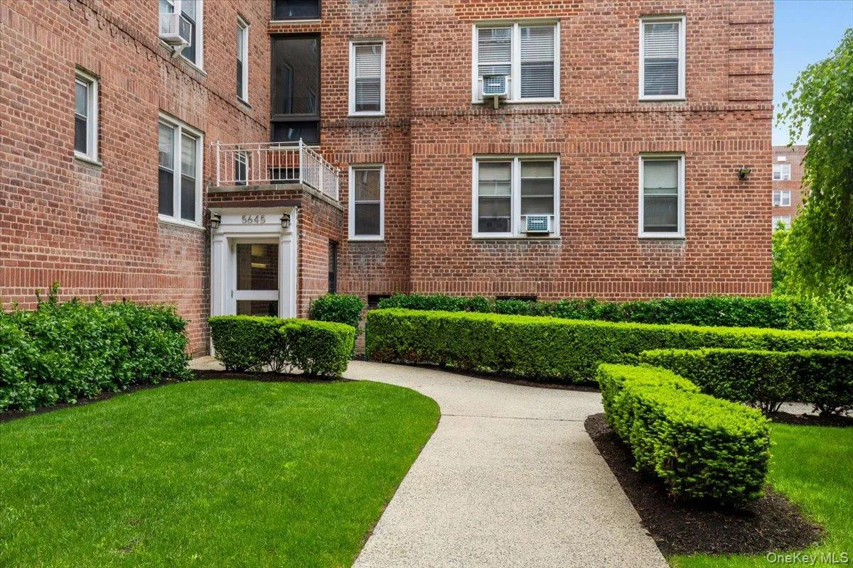 Brick facade building featuring a prominent entrance with white trim, a second-story balcony with white railing, a concrete pathway, manicured lawns, and shaped hedges Brick facade building featuring a prominent entrance with white trim, a second-story balcony with white railing, a concrete pathway, manicured lawns, and shaped hedges