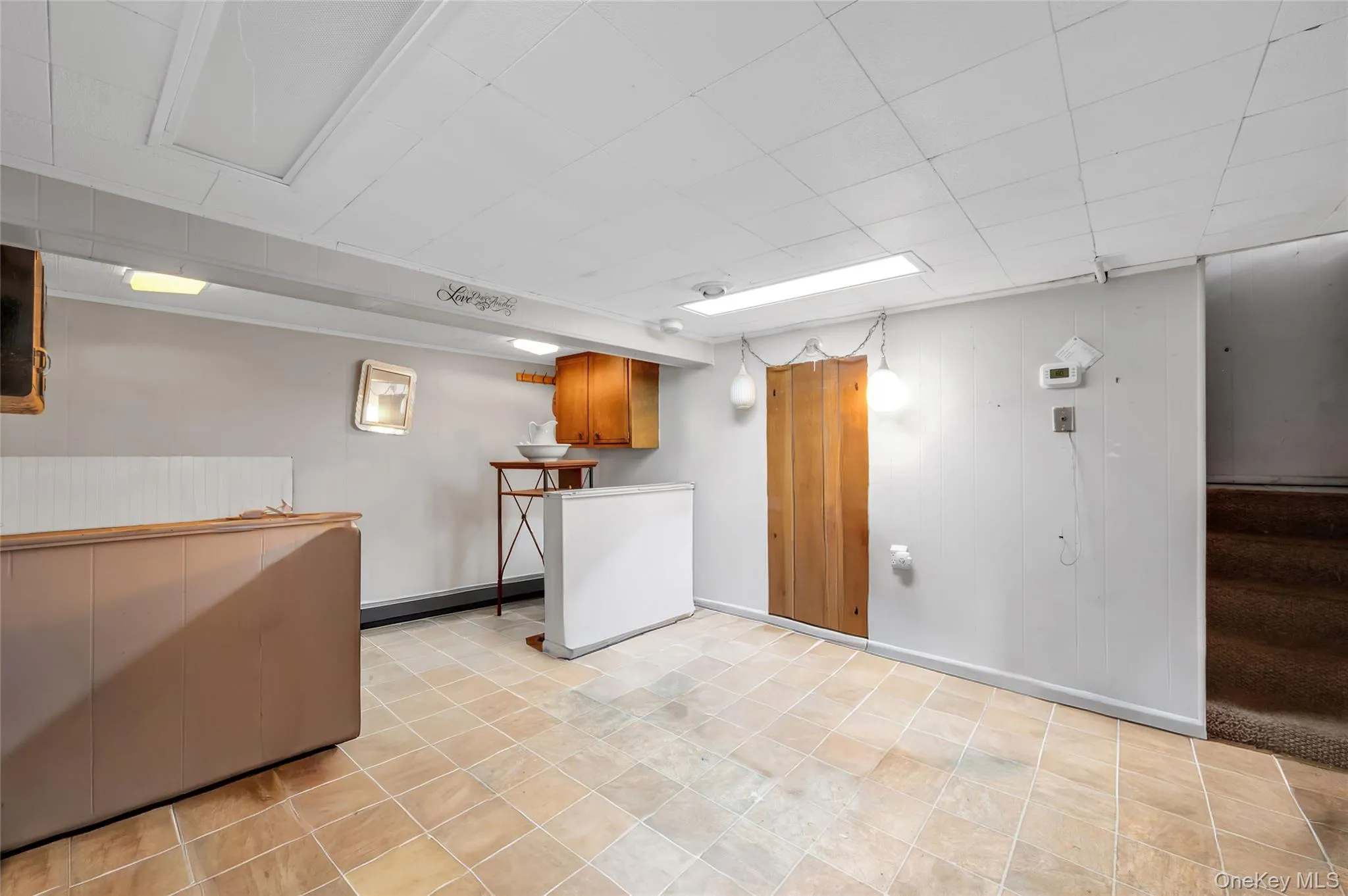 Finished lower level space featuring square tile flooring, drop ceiling with fluorescent lighting, and a built-in wood cabinet Finished lower level space featuring square tile flooring, drop ceiling with fluorescent lighting, and a built-in wood cabinet