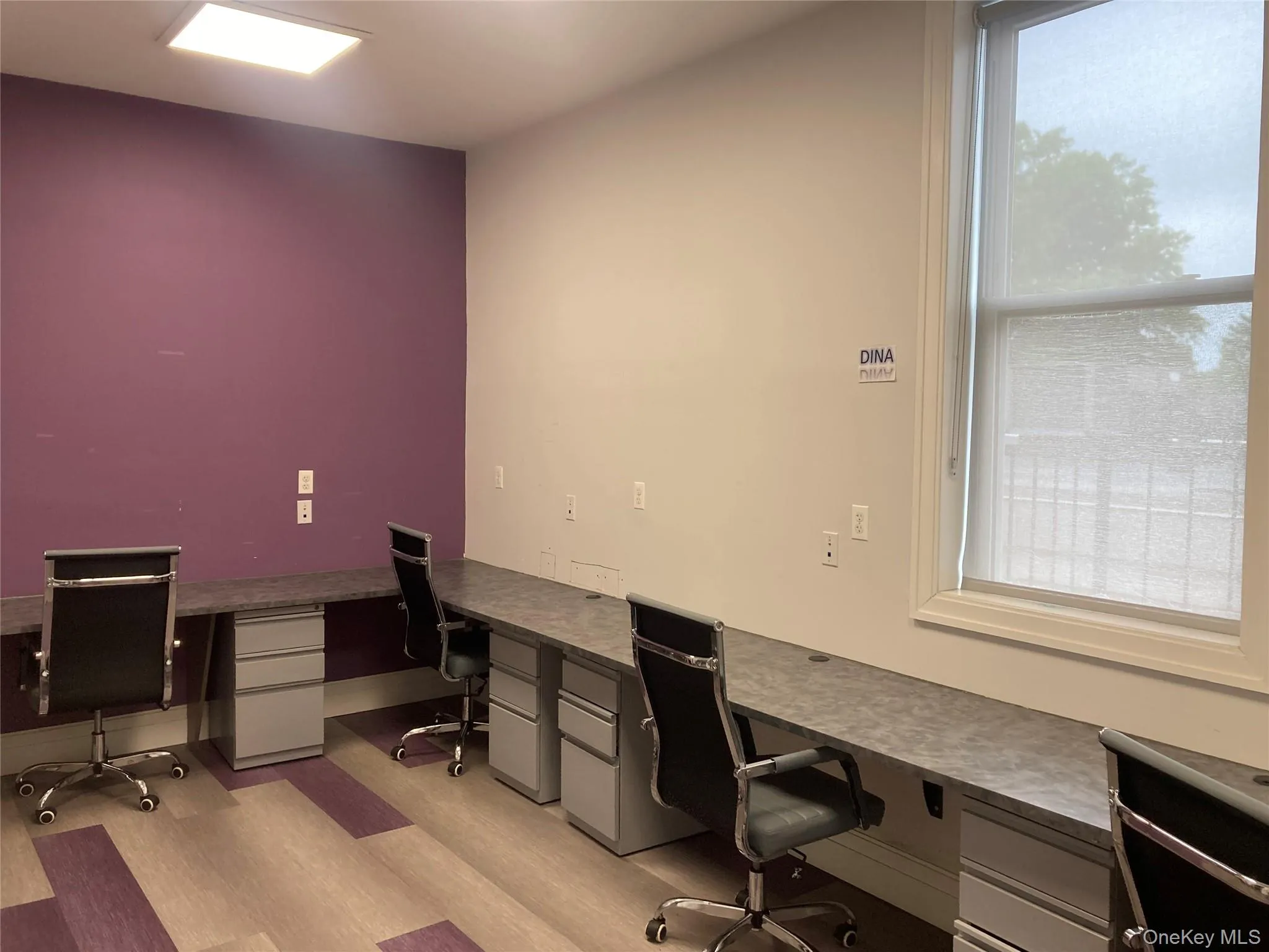 Dedicated workspace featuring a large window with roller shade, integrated desk units, and patterned wood-finish flooring Dedicated workspace featuring a large window with roller shade, integrated desk units, and patterned wood-finish flooring