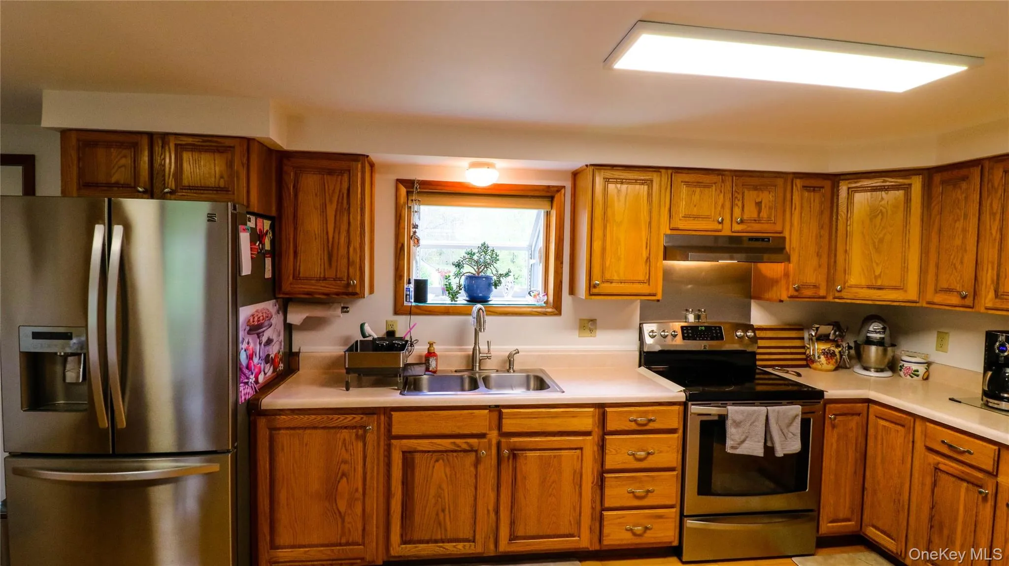 Kitchen featuring wood cabinetry, light-toned countertops, and stainless steel appliances including a refrigerator, range, and hood Kitchen featuring wood cabinetry, light-toned countertops, and stainless steel appliances including a refrigerator, range, and hood