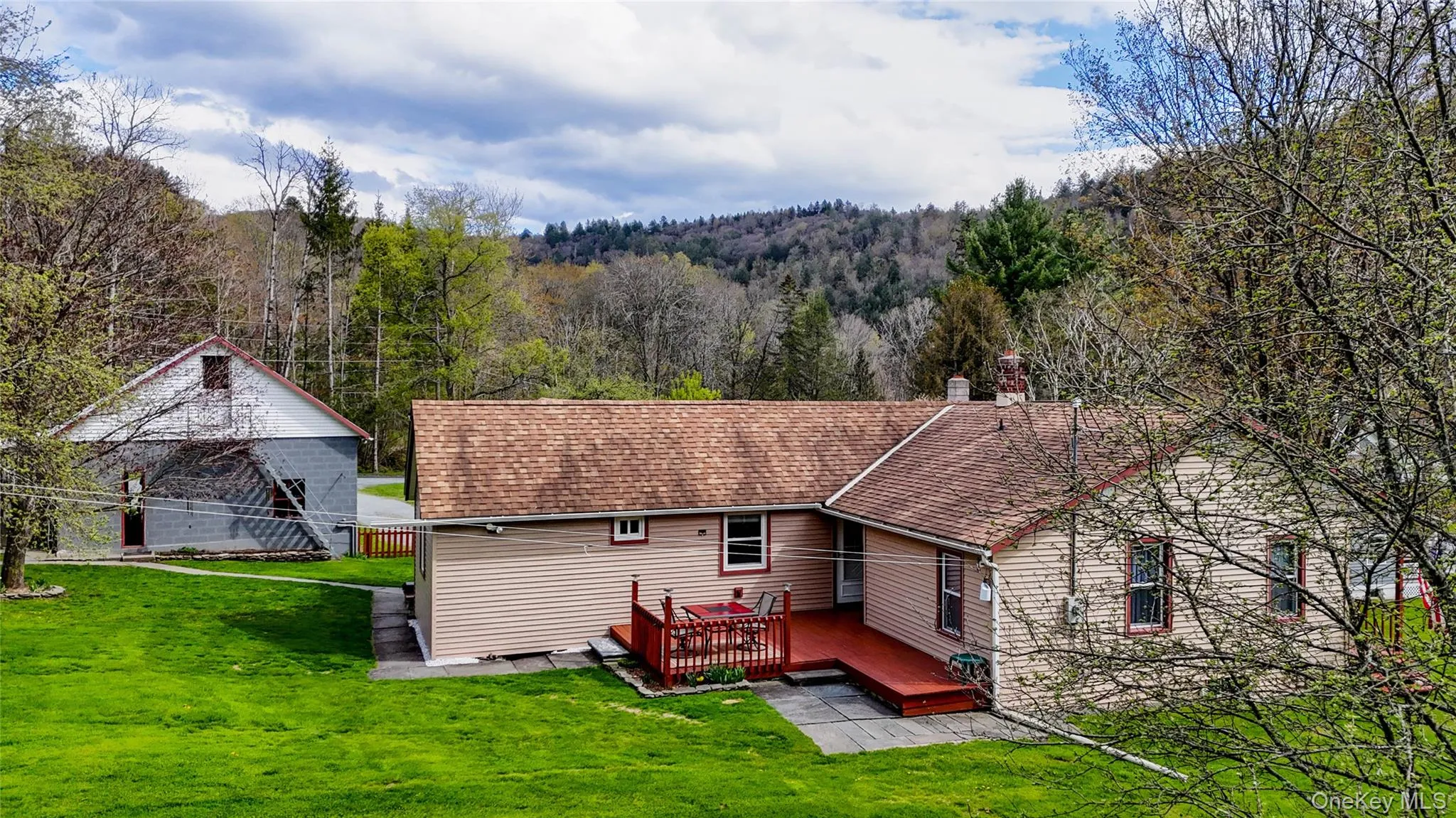 Expansive green lawn with a tan siding residence featuring a red wood-finish deck and a separate two-story barn with a light gray and white exterior Expansive green lawn with a tan siding residence featuring a red wood-finish deck and a separate two-story barn with a light gray and white exterior