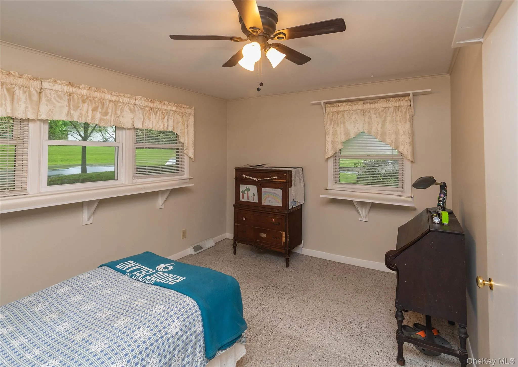 Neutral wall paint, two windows with blinds, ceiling fan with light fixture, light-toned carpet, and white baseboards Neutral wall paint, two windows with blinds, ceiling fan with light fixture, light-toned carpet, and white baseboards