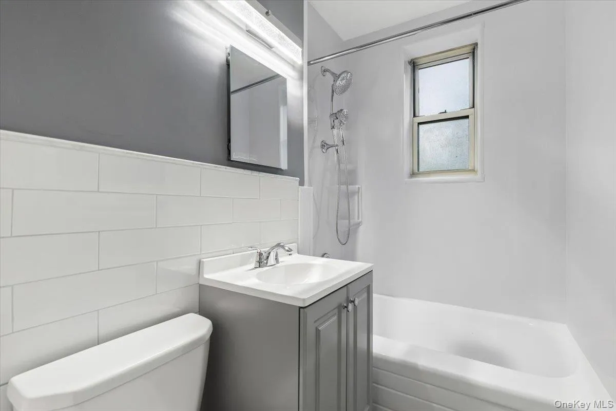 Bathroom featuring a white subway tile wainscoting, a gray vanity with a white countertop and integrated sink, a toilet, a shower/tub combination with a chrome shower fixture, and a window Bathroom featuring a white subway tile wainscoting, a gray vanity with a white countertop and integrated sink, a toilet, a shower/tub combination with a chrome shower fixture, and a window