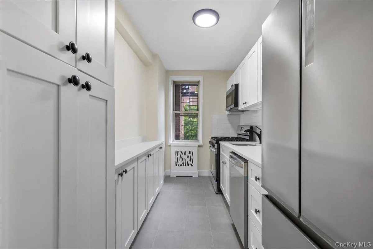 Galley kitchen featuring white cabinetry, stainless steel appliances, a window with exterior views, light-toned countertops, and grey tile flooring Galley kitchen featuring white cabinetry, stainless steel appliances, a window with exterior views, light-toned countertops, and grey tile flooring