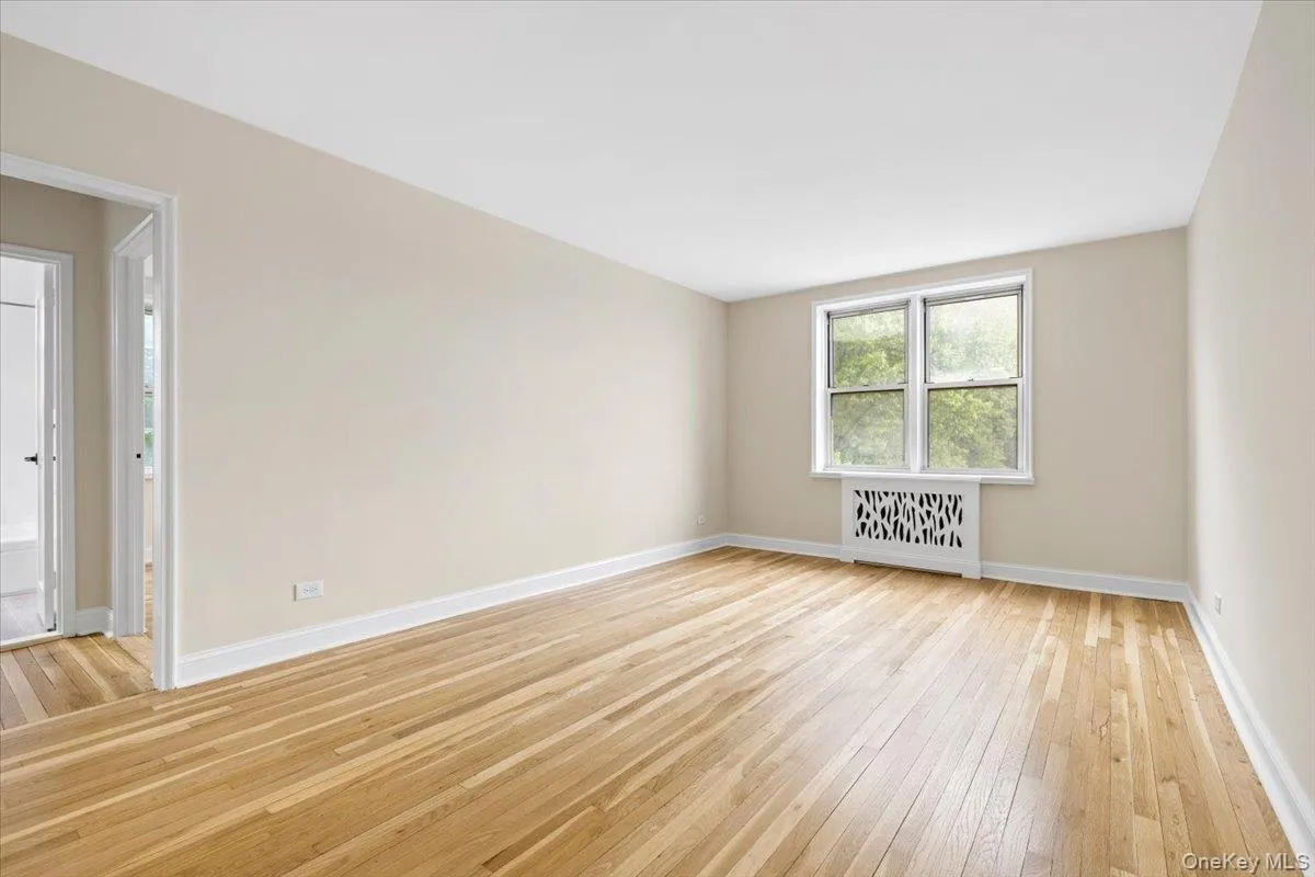 Spacious living room featuring wood-finish flooring, neutral-toned walls, white trim, a large window, and a decorative radiator cover Spacious living room featuring wood-finish flooring, neutral-toned walls, white trim, a large window, and a decorative radiator cover