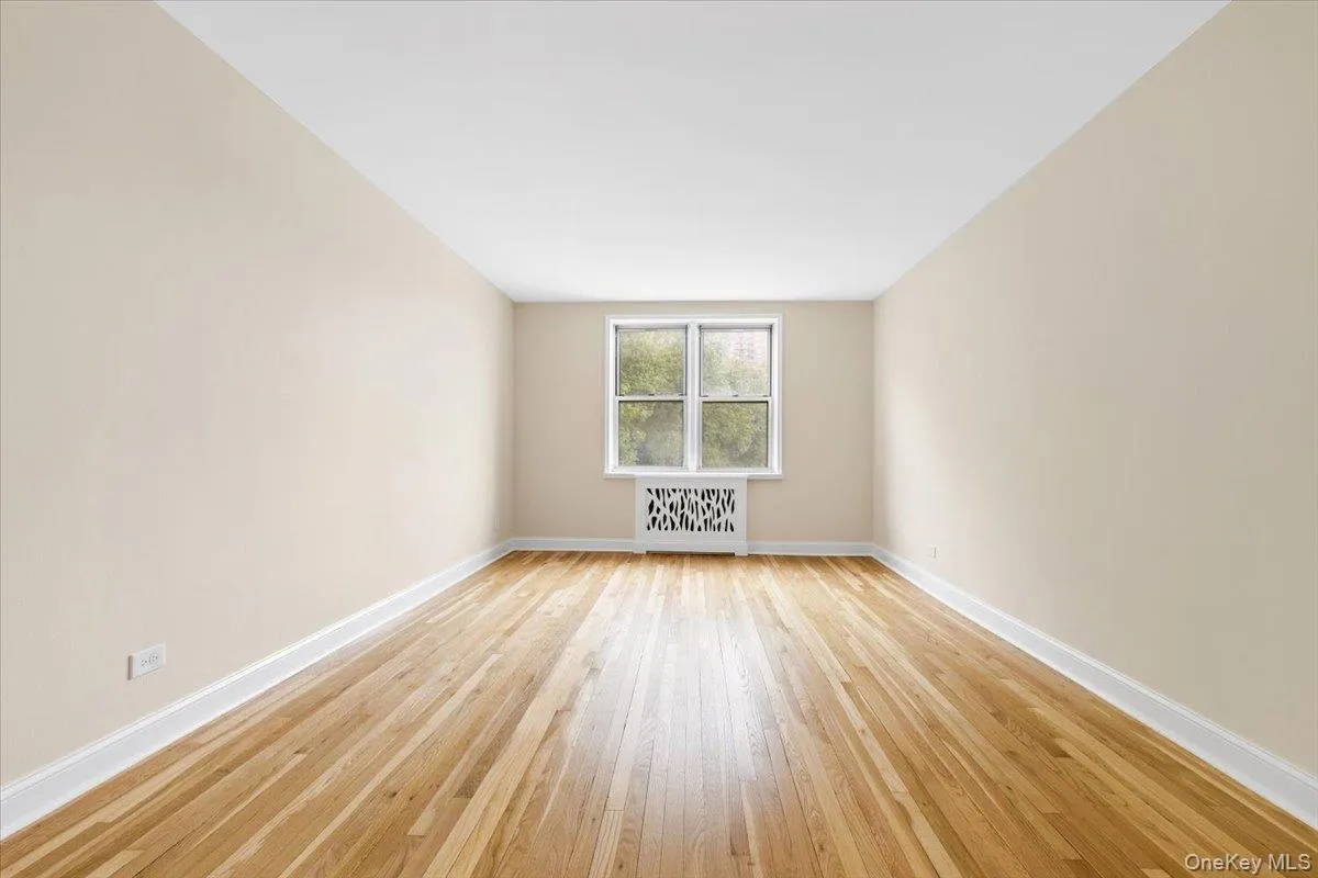 Bright interior living room featuring wood-finish flooring, light-toned wall paint, crisp white baseboards, a multi-pane window, and a decorative radiator cover Bright interior living room featuring wood-finish flooring, light-toned wall paint, crisp white baseboards, a multi-pane window, and a decorative radiator cover