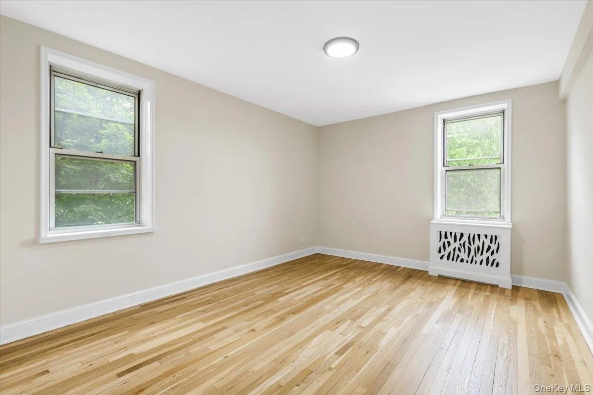 Spacious bedroom featuring light wood-finish flooring, neutral wall tones, and two operable windows Spacious bedroom featuring light wood-finish flooring, neutral wall tones, and two operable windows