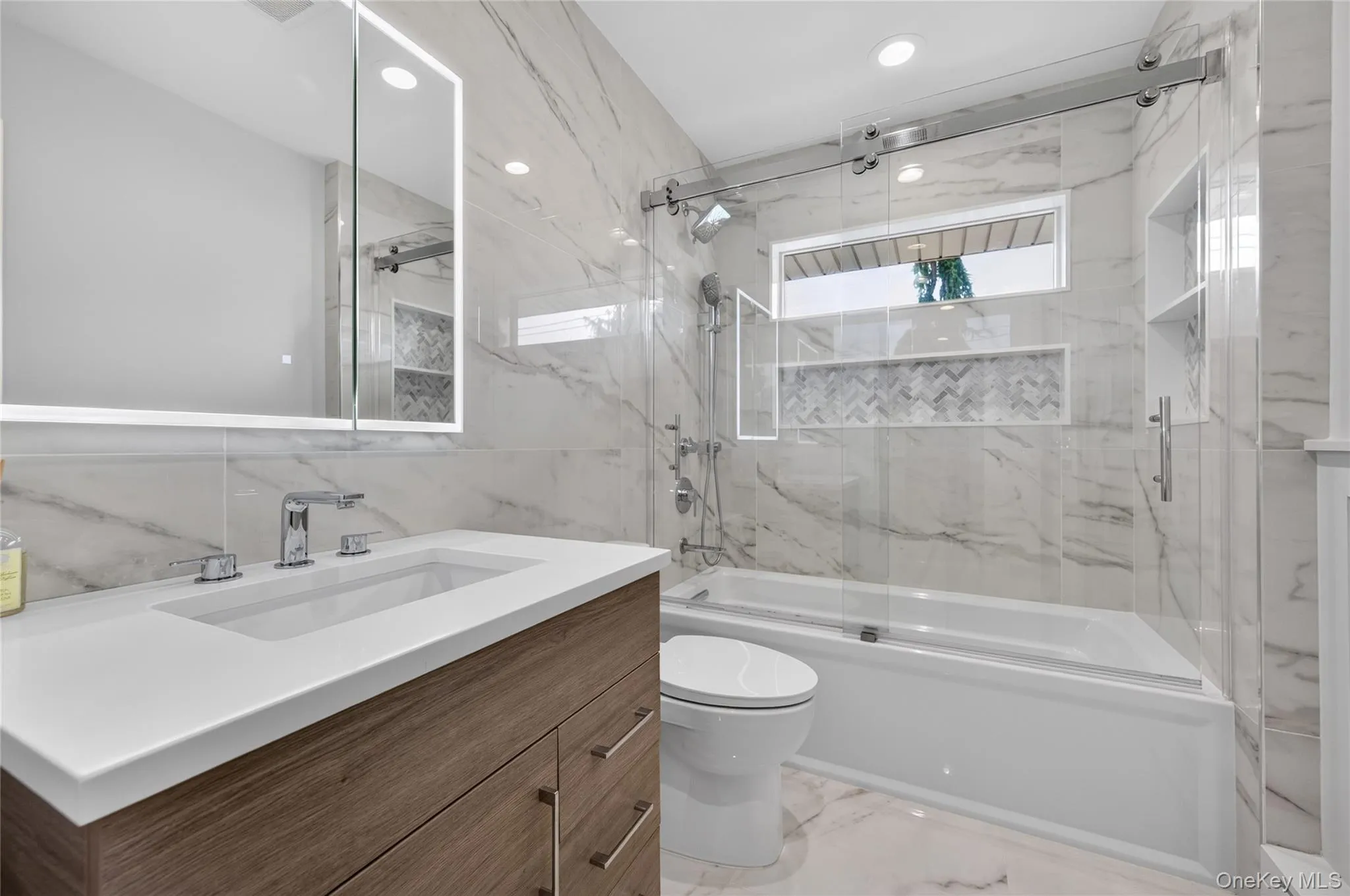 Hall bathroom featuring a wood-finish vanity with an integrated sink and chrome faucet, an illuminated mirror, a tub with a sliding glass door enclosure with built-in niche Hall bathroom featuring a wood-finish vanity with an integrated sink and chrome faucet, an illuminated mirror, a tub with a sliding glass door enclosure with built-in niche