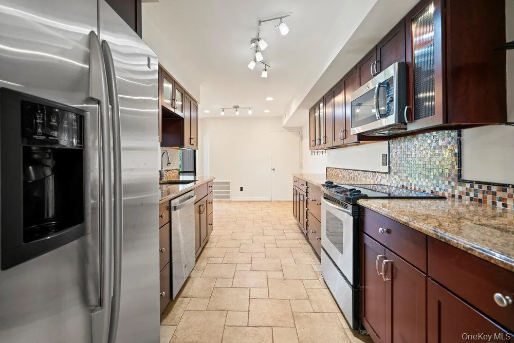 Galley kitchen featuring stainless steel appliances, dark wood cabinetry, granite countertops, and mosaic tile backsplash Galley kitchen featuring stainless steel appliances, dark wood cabinetry, granite countertops, and mosaic tile backsplash