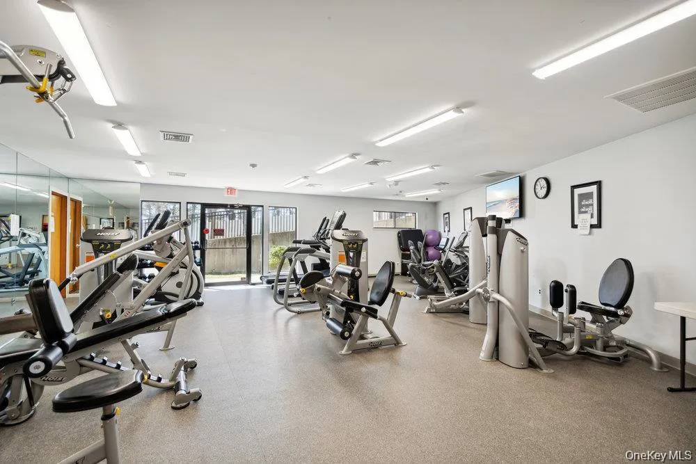 Fitness center featuring resilient rubber flooring, mirrored wall panels, fluorescent overhead lighting, and extensive workout equipment Fitness center featuring resilient rubber flooring, mirrored wall panels, fluorescent overhead lighting, and extensive workout equipment