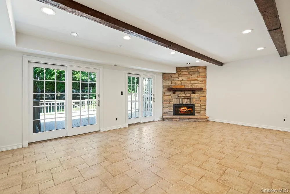 Spacious room featuring tile flooring, a corner stone-clad fireplace with a wood mantel, and exposed wooden ceiling beams Spacious room featuring tile flooring, a corner stone-clad fireplace with a wood mantel, and exposed wooden ceiling beams
