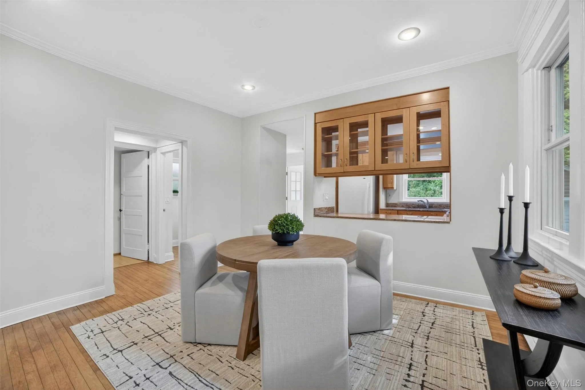Open-concept living area with wood-finish flooring, recessed lighting, crown molding, and a pass-through window with glass-front wood cabinetry Open-concept living area with wood-finish flooring, recessed lighting, crown molding, and a pass-through window with glass-front wood cabinetry