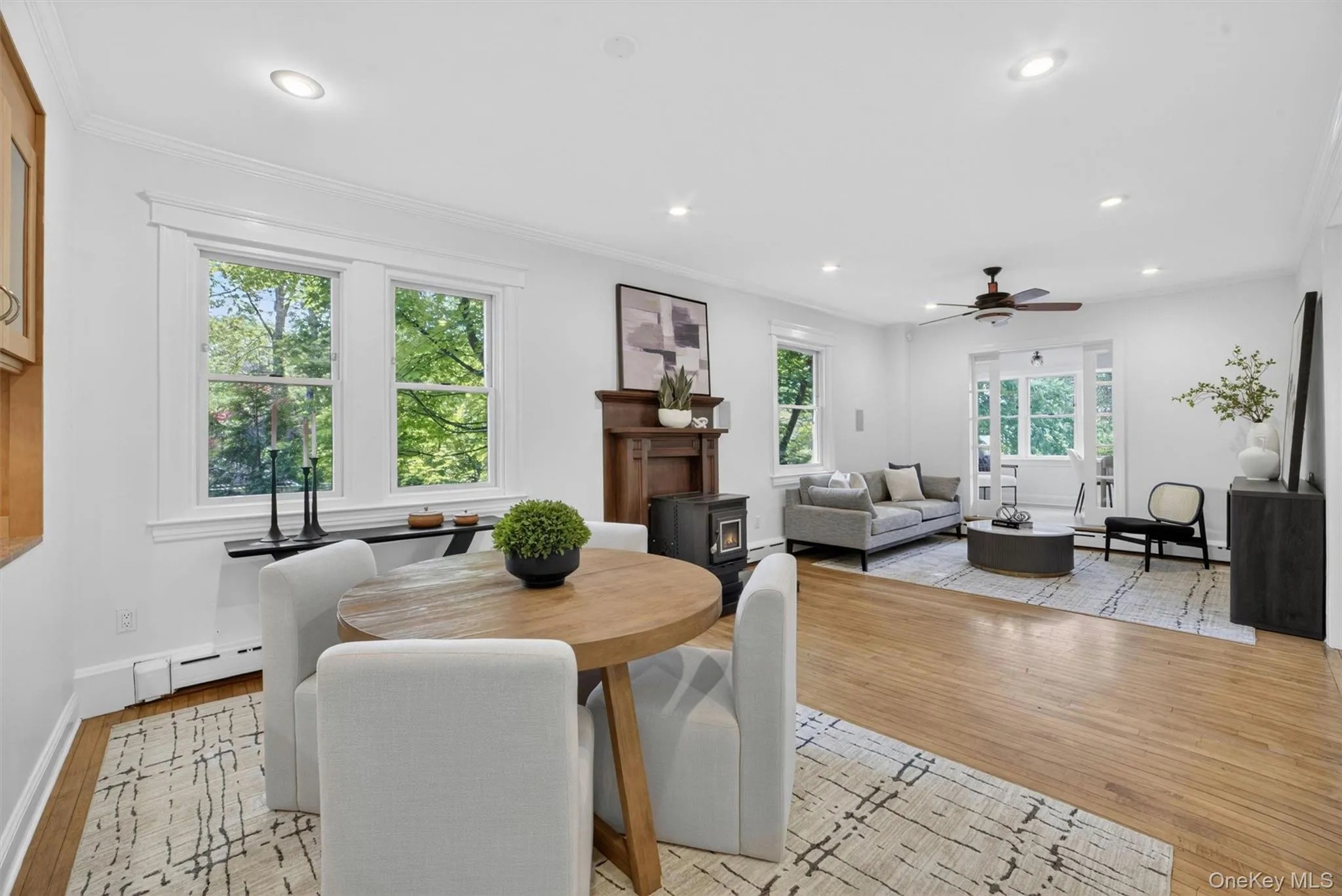 Hardwood flooring in a bright open-concept living area featuring recessed lighting, a decorative wooden fireplace surround with a hearth, and multiple windows Hardwood flooring in a bright open-concept living area featuring recessed lighting, a decorative wooden fireplace surround with a hearth, and multiple windows