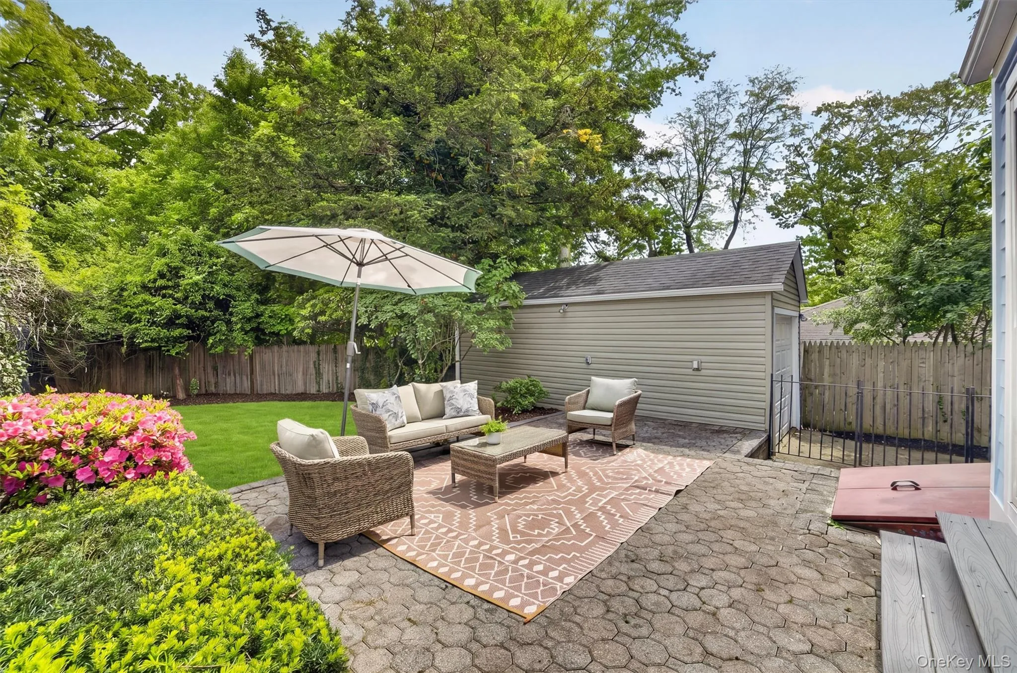 Paver patio with a separate detached garage structure, lush green lawn, mature trees, and established flowering shrubs Paver patio with a separate detached garage structure, lush green lawn, mature trees, and established flowering shrubs
