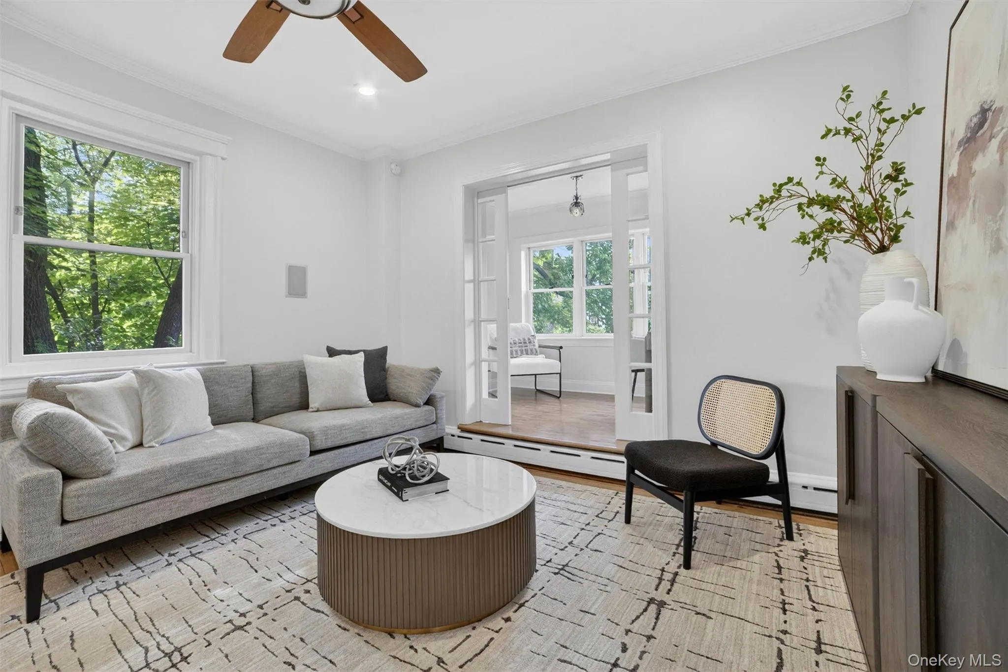 Living area with white walls and crown molding, featuring a ceiling fan with wood-finish blades, a large window, and French doors opening to an additional room Living area with white walls and crown molding, featuring a ceiling fan with wood-finish blades, a large window, and French doors opening to an additional room