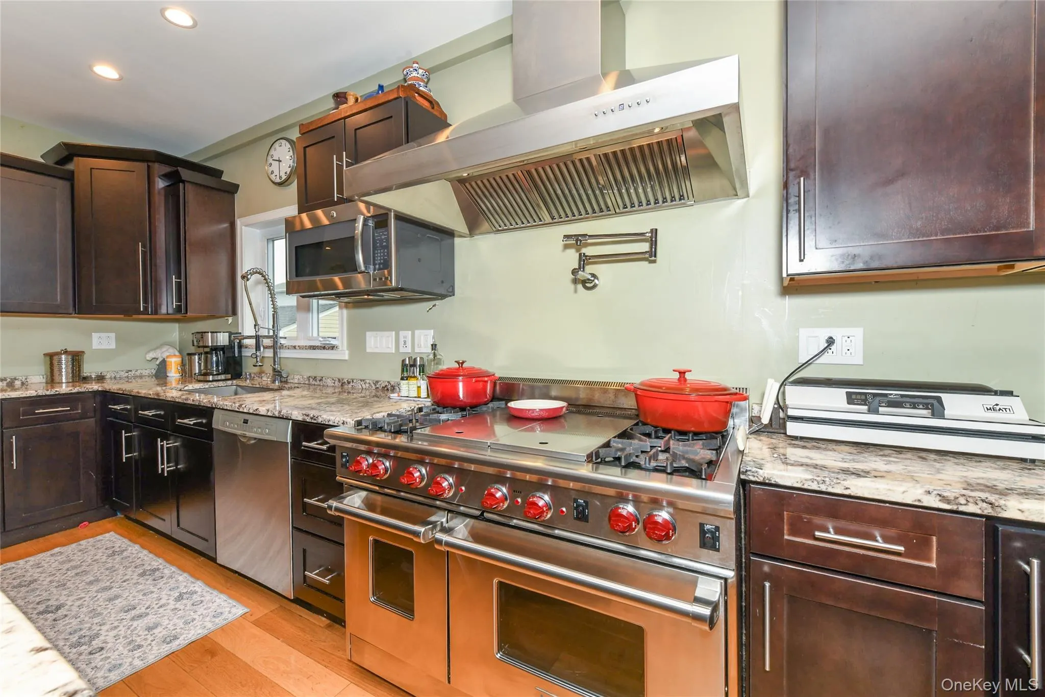Kitchen with stainless steel appliances, wall chimney range hood, dark brown cabinetry, light stone countertops, and light wood-style floors Kitchen with stainless steel appliances, wall chimney range hood, dark brown cabinetry, light stone countertops, and light wood-style floors