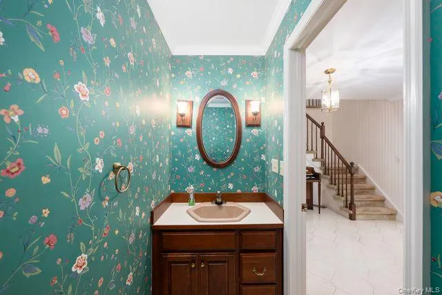 Powder room, 1st floor, featuring floral wallpaper, a dark wood vanity with a light countertop, an oval mirror, and wall-mounted sconces Powder room, 1st floor, featuring floral wallpaper, a dark wood vanity with a light countertop, an oval mirror, and wall-mounted sconces