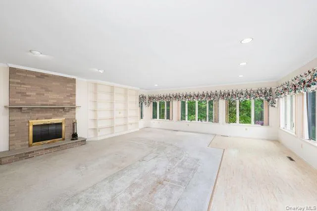 Spacious interior sun filled room featuring a brick fireplace with a brass-trimmed firebox, extensive built-in shelving, a wall of windows, recessed lighting, and a combination of carpet and light finish flooring Spacious interior sun filled room featuring a brick fireplace with a brass-trimmed firebox, extensive built-in shelving, a wall of windows, recessed lighting, and a combination of carpet and light finish flooring