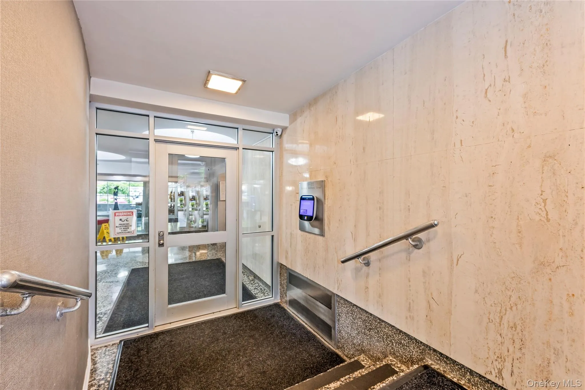 Building entrance featuring a glass door with an aluminum frame, an adjacent biometric access control system, and a wall of light-toned stone tiling Building entrance featuring a glass door with an aluminum frame, an adjacent biometric access control system, and a wall of light-toned stone tiling