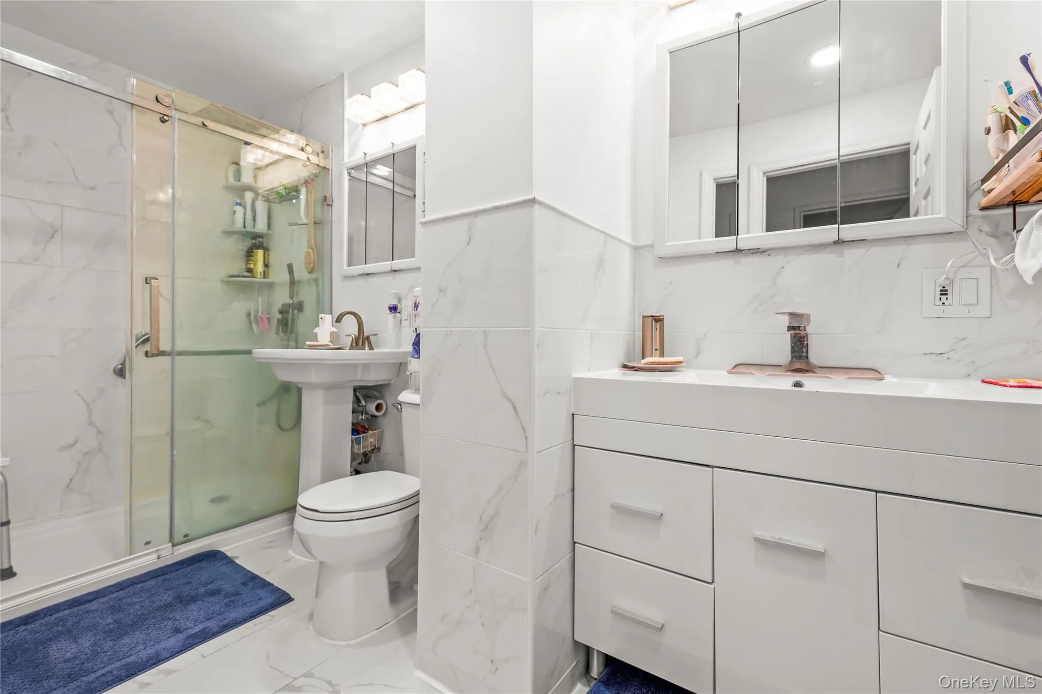 Bathroom featuring a glass-enclosed shower, white marble-look wall and floor tiling, a pedestal sink, and a white vanity with an integrated sink and storage Bathroom featuring a glass-enclosed shower, white marble-look wall and floor tiling, a pedestal sink, and a white vanity with an integrated sink and storage