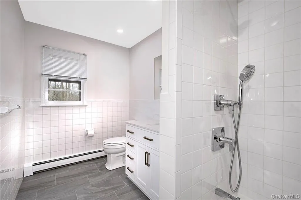 Full bathroom featuring white subway tile, a white vanity with dark hardware, a walk-in shower with a handheld showerhead, and wood-finish tile flooring Full bathroom featuring white subway tile, a white vanity with dark hardware, a walk-in shower with a handheld showerhead, and wood-finish tile flooring