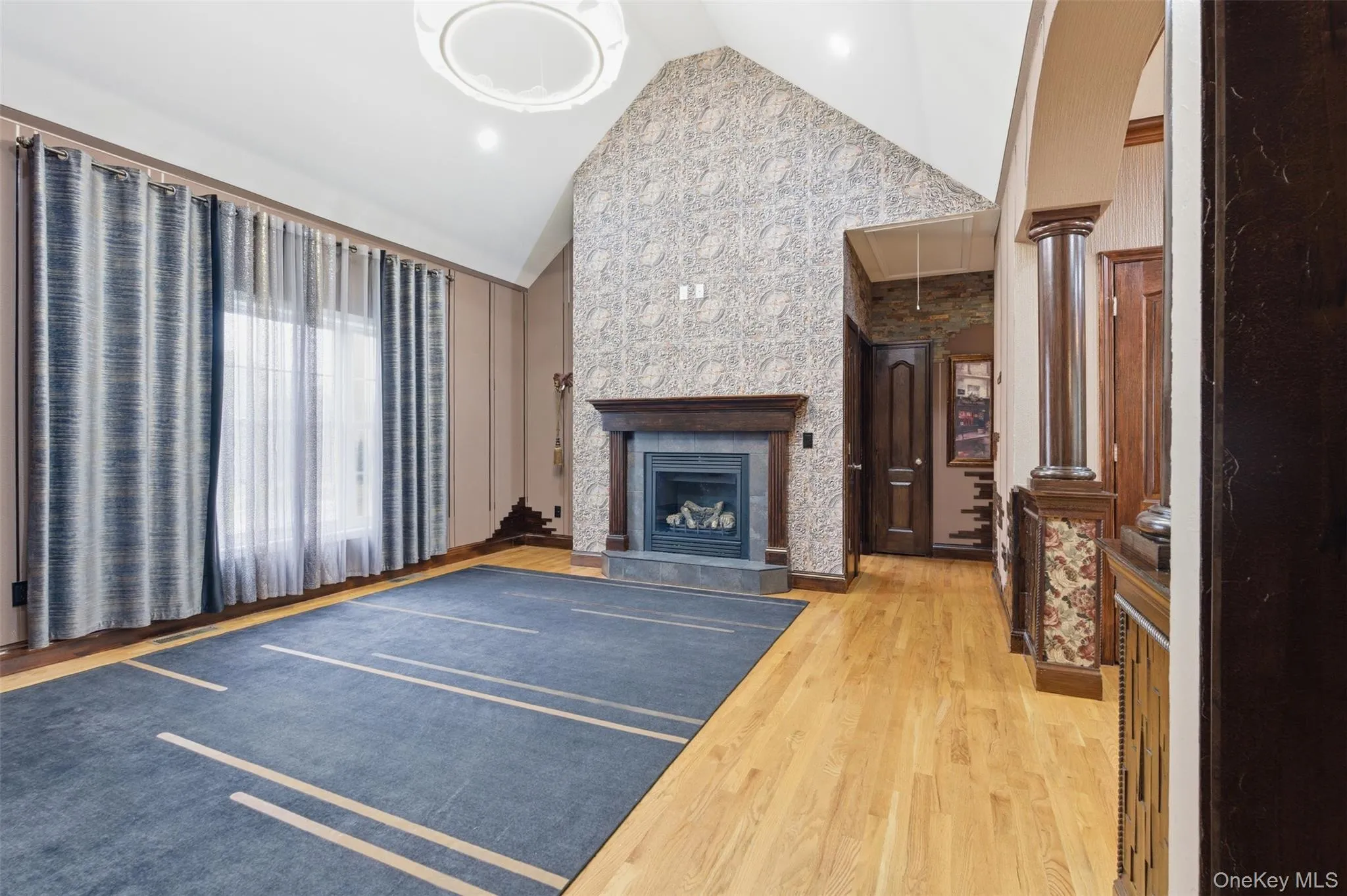 Vaulted ceiling with an elegant circular light fixture, hardwood flooring, and a fireplace featuring a dark wood mantle and stone surround Vaulted ceiling with an elegant circular light fixture, hardwood flooring, and a fireplace featuring a dark wood mantle and stone surround