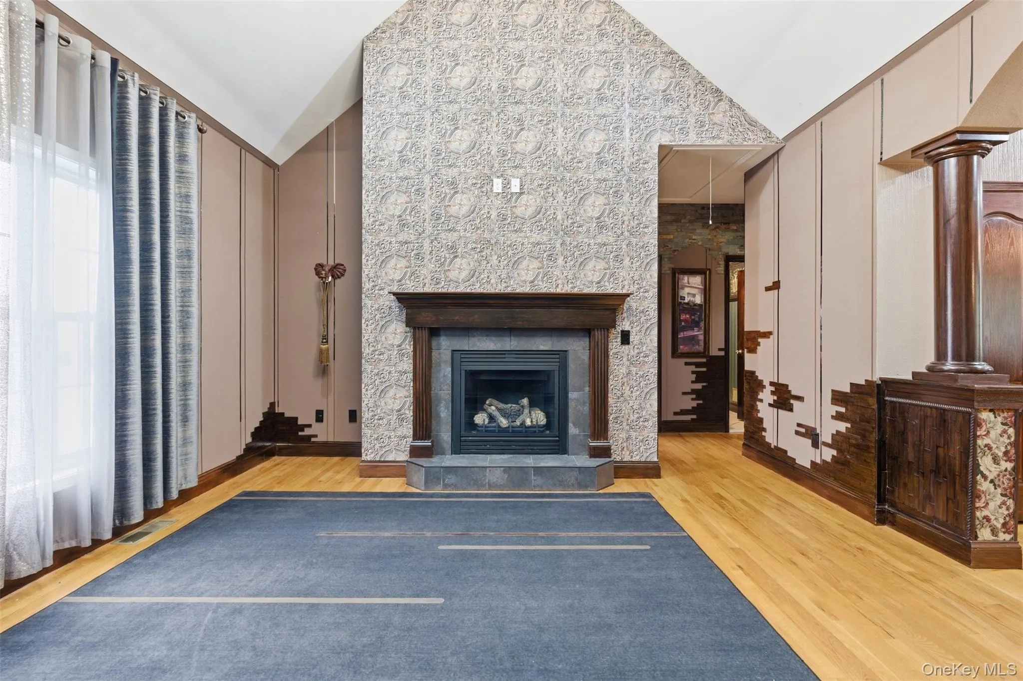 Vaulted ceiling room featuring a gas fireplace with a dark wood mantel and slate hearth, patterned wall paneling, light wood-finish flooring, and a column with decorative wood detailing Vaulted ceiling room featuring a gas fireplace with a dark wood mantel and slate hearth, patterned wall paneling, light wood-finish flooring, and a column with decorative wood detailing