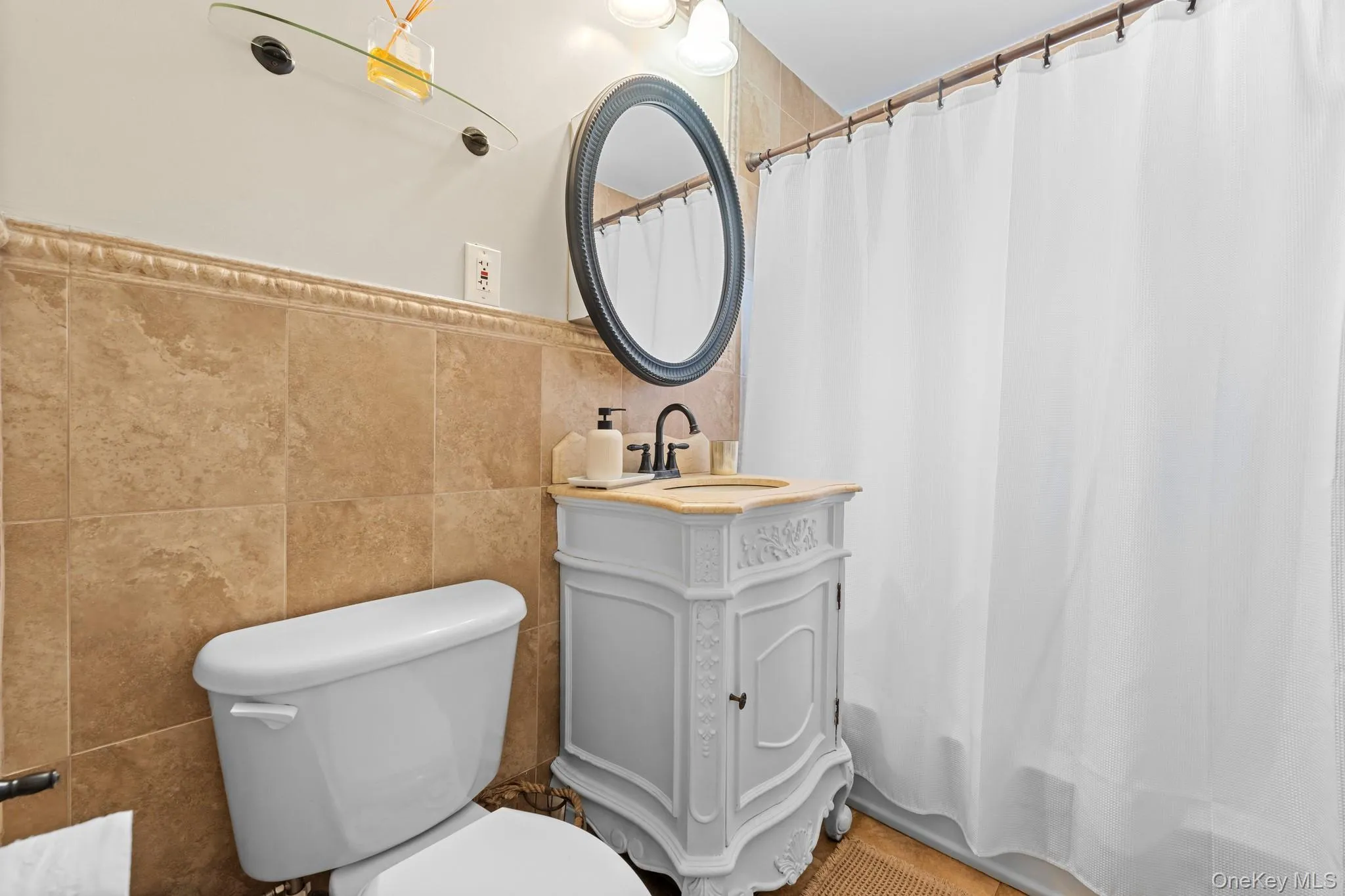 Bathroom featuring a white vanity with an undermount sink, an oval mirror, a glass shelf, and a white toilet Bathroom featuring a white vanity with an undermount sink, an oval mirror, a glass shelf, and a white toilet