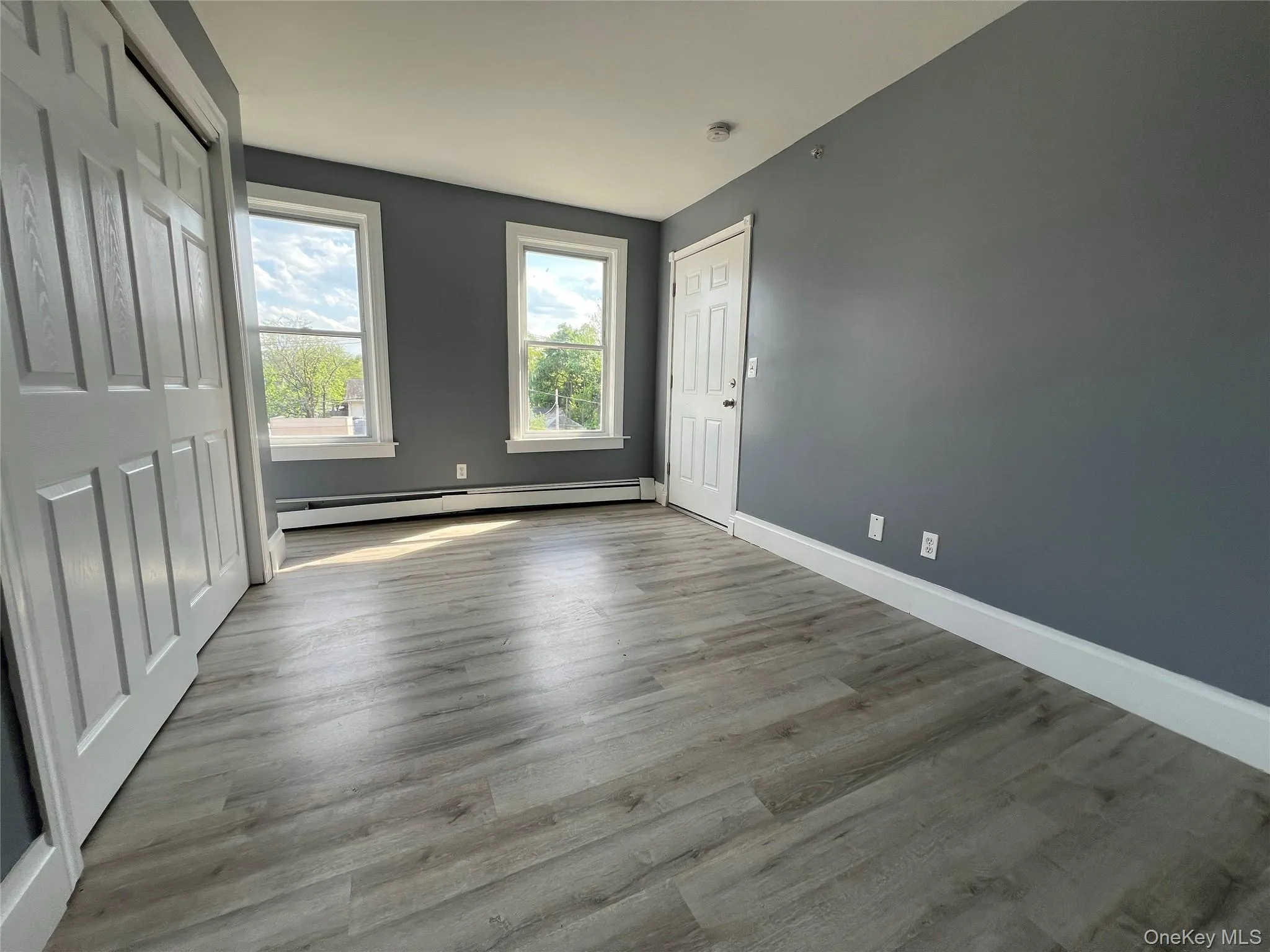 Spacious room featuring wood-finish flooring, two windows with white trim, baseboard heating, and a solid white door Spacious room featuring wood-finish flooring, two windows with white trim, baseboard heating, and a solid white door