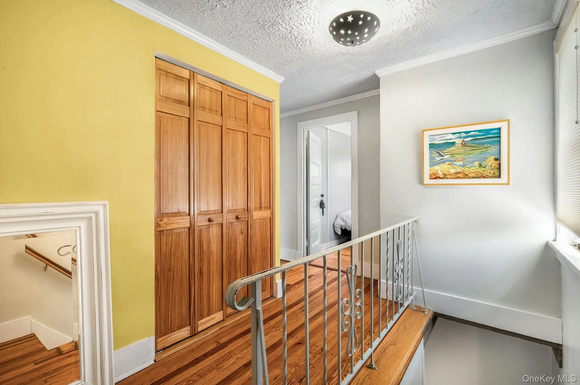 Hallway with wood-finish flooring, bi-fold closet doors, a decorative ceiling light fixture, and a metal railing Hallway with wood-finish flooring, bi-fold closet doors, a decorative ceiling light fixture, and a metal railing