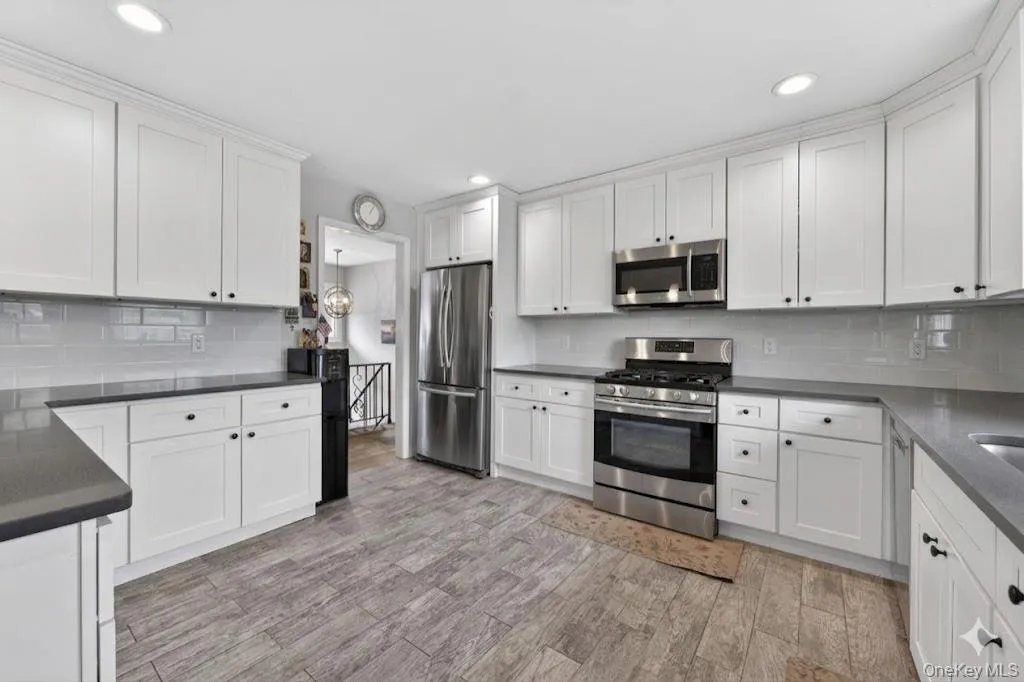 Modern kitchen featuring white shaker cabinetry, dark countertops, a subway tile backsplash, stainless steel appliances, and wood-look plank flooring Modern kitchen featuring white shaker cabinetry, dark countertops, a subway tile backsplash, stainless steel appliances, and wood-look plank flooring