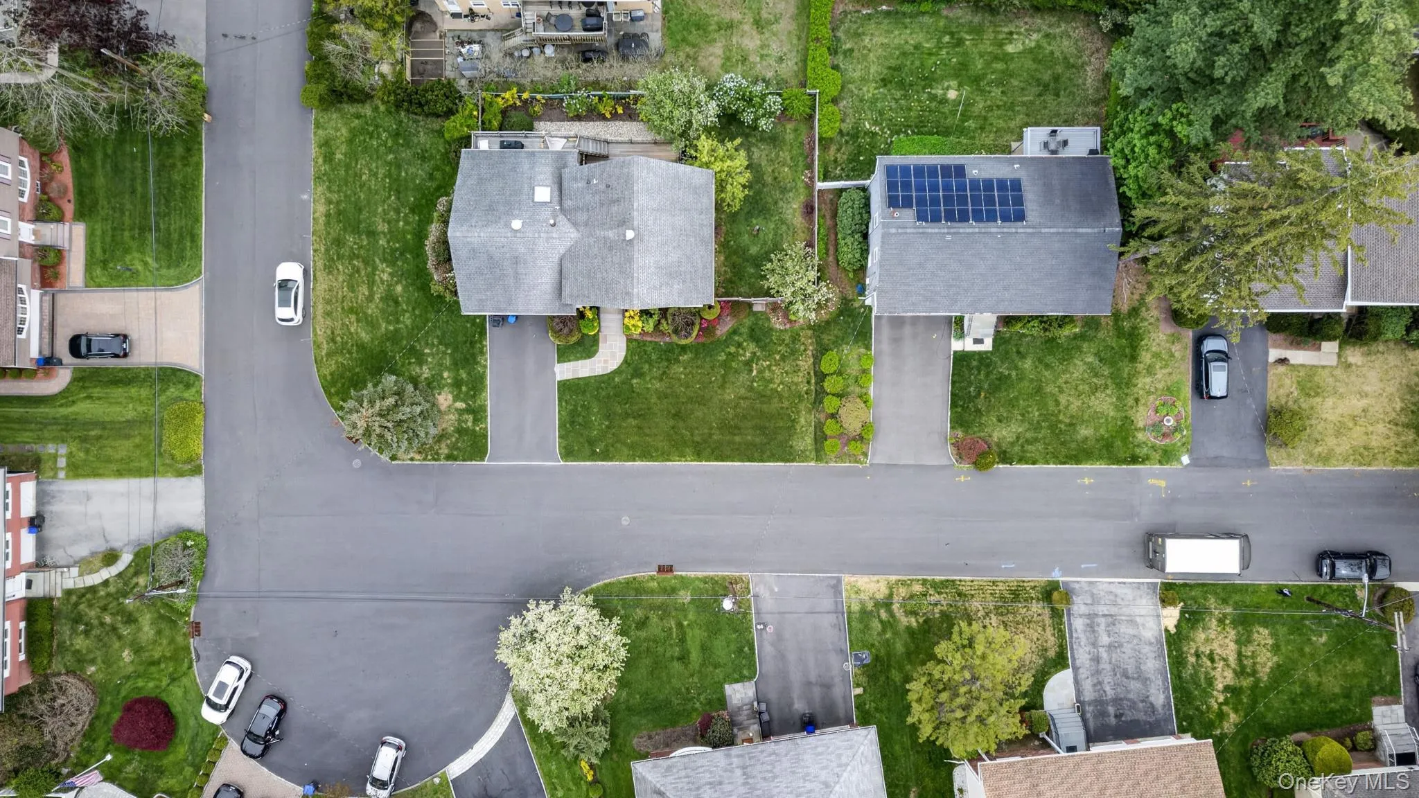 Residential street featuring asphalt paving, concrete sidewalks, and properties with pitched shingle roofs, green lawns, and mature trees and cul-de-sac locations Residential street featuring asphalt paving, concrete sidewalks, and properties with pitched shingle roofs, green lawns, and mature trees and cul-de-sac locations