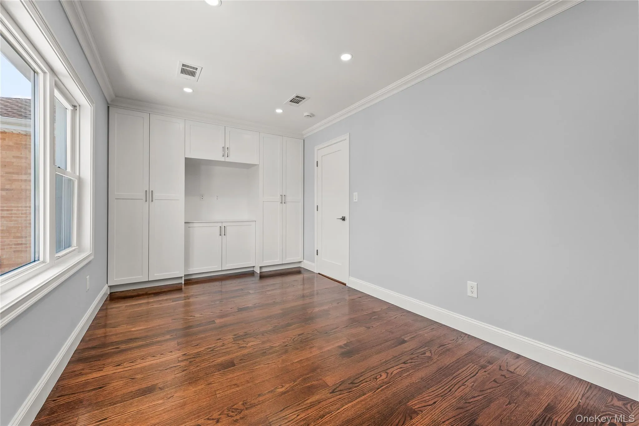 Interior room featuring custom extensive built-in white cabinetry, lustrous wood-finish flooring, recessed lighting, double-hung windows, and crown molding Interior room featuring custom extensive built-in white cabinetry, lustrous wood-finish flooring, recessed lighting, double-hung windows, and crown molding