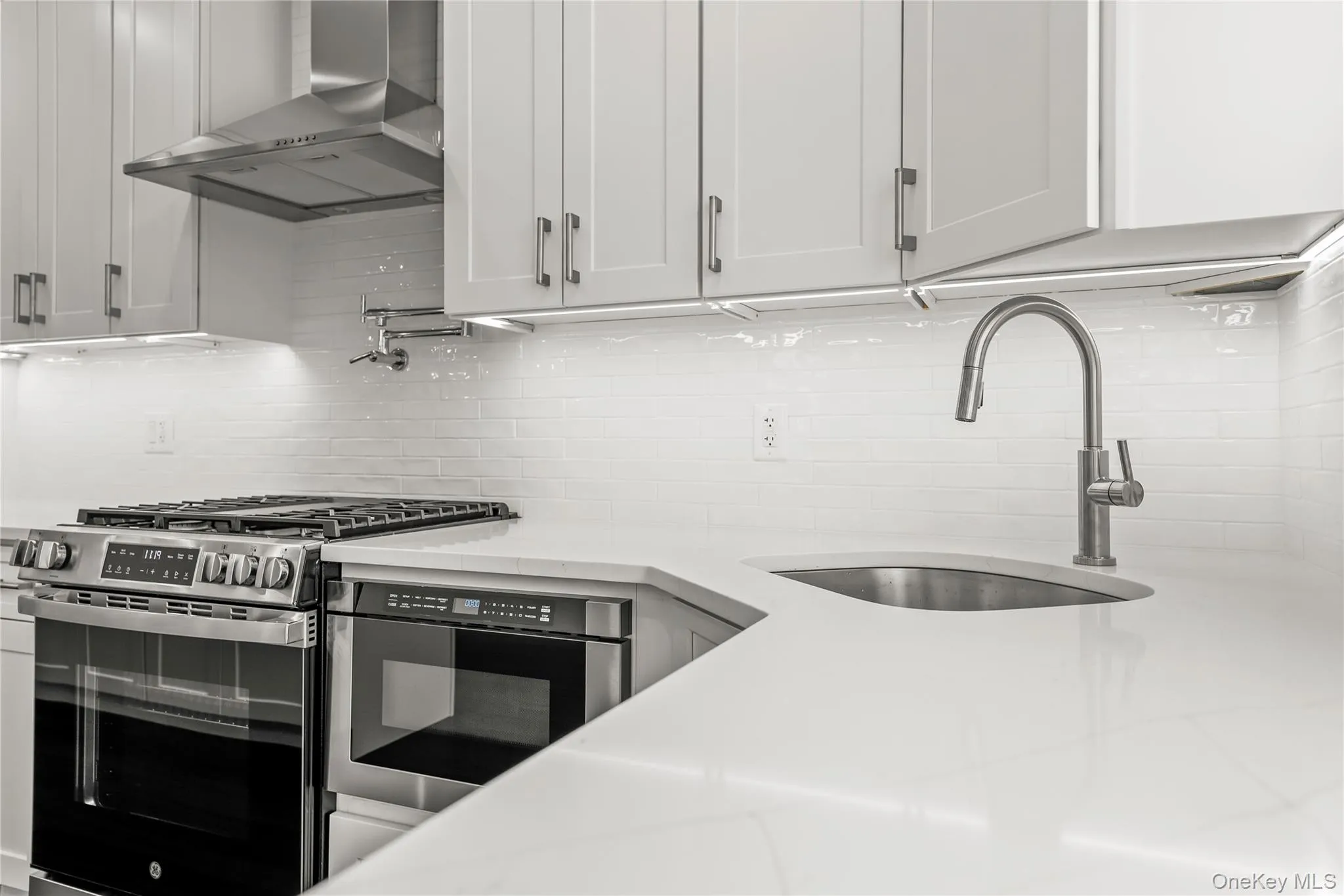 White cabinetry with brushed nickel hardware, stainless steel range with overhead hood, integrated microwave, white subway tile backsplash, and undermount stainless steel sink with gooseneck faucet White cabinetry with brushed nickel hardware, stainless steel range with overhead hood, integrated microwave, white subway tile backsplash, and undermount stainless steel sink with gooseneck faucet