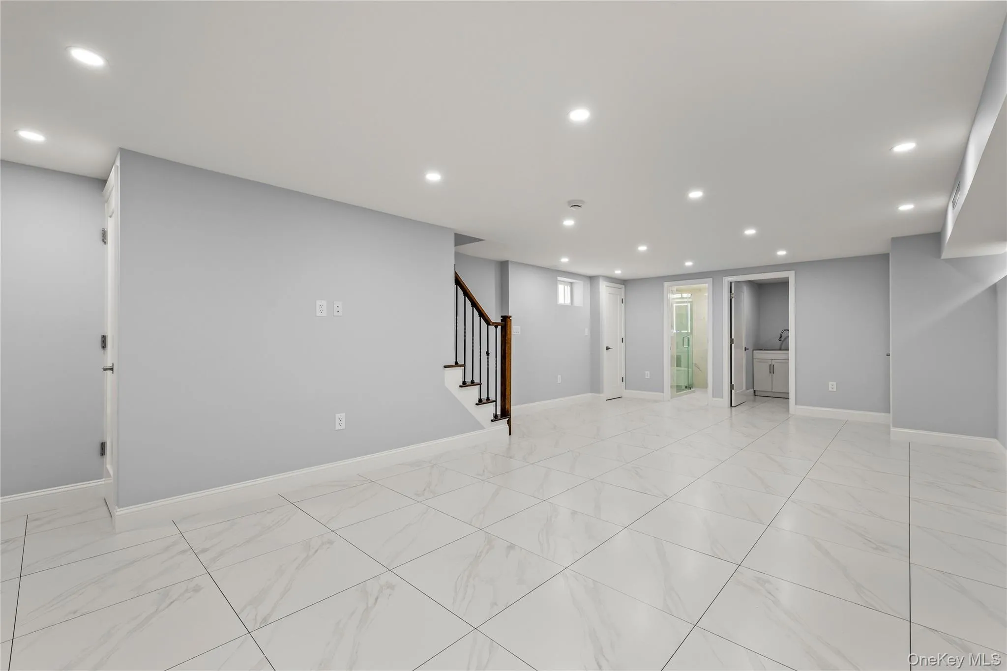 Spacious room featuring large format tile flooring, recessed lighting, a modern staircase with dark wood handrail and wrought iron balusters, and light gray wall finishes Spacious room featuring large format tile flooring, recessed lighting, a modern staircase with dark wood handrail and wrought iron balusters, and light gray wall finishes