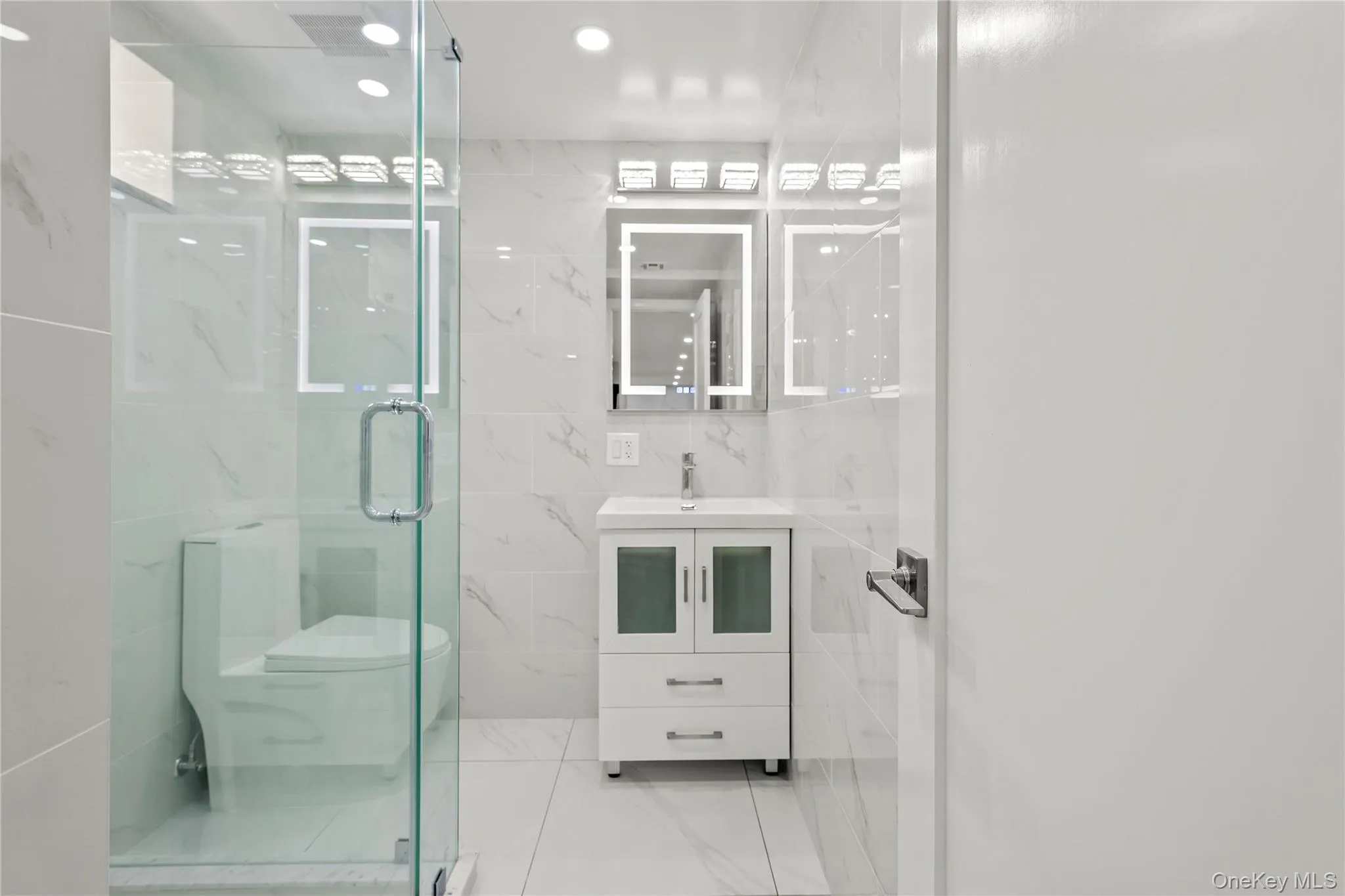 Bathroom featuring a glass-enclosed shower, a white vanity with a rectangular sink, an illuminated mirror, and marble-look wall tiles Bathroom featuring a glass-enclosed shower, a white vanity with a rectangular sink, an illuminated mirror, and marble-look wall tiles
