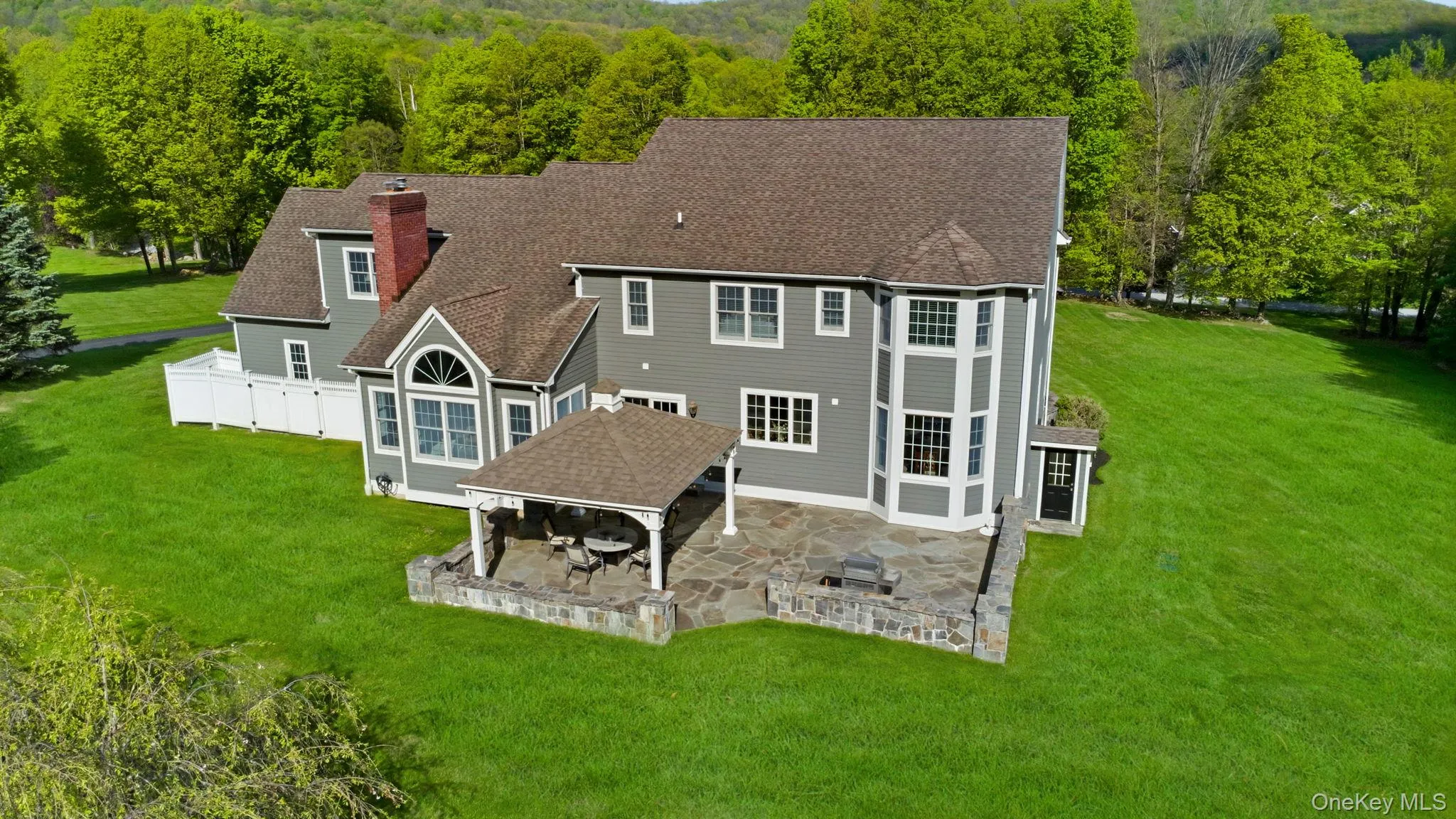 Gray siding residence featuring a covered flagstone patio, stone retaining walls, multiple windows, and a prominent red brick chimney, set amidst extensive green lawns Gray siding residence featuring a covered flagstone patio, stone retaining walls, multiple windows, and a prominent red brick chimney, set amidst extensive green lawns
