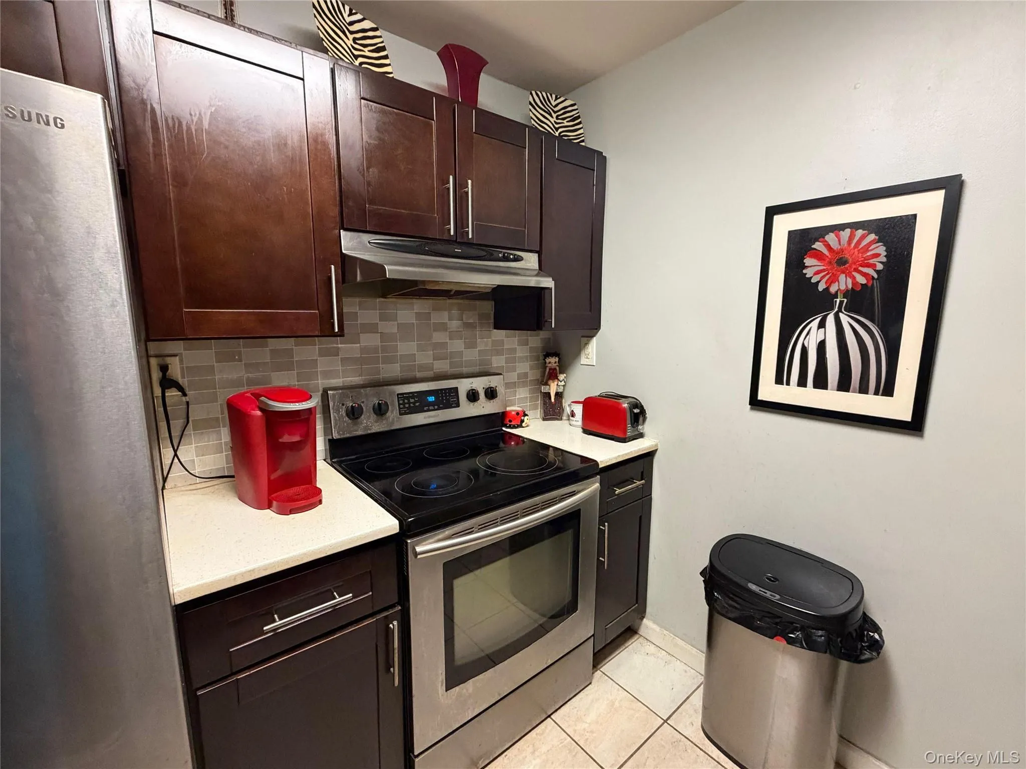 Compact kitchen featuring dark wood cabinetry, stainless steel appliances, a tiled backsplash, and light-toned countertops Compact kitchen featuring dark wood cabinetry, stainless steel appliances, a tiled backsplash, and light-toned countertops