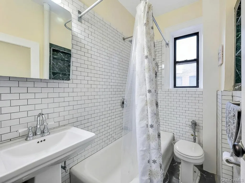 Bathroom featuring white subway tile walls, a pedestal sink with chrome faucet, a standard bathtub, a window with black trim, and dark stone flooring Bathroom featuring white subway tile walls, a pedestal sink with chrome faucet, a standard bathtub, a window with black trim, and dark stone flooring