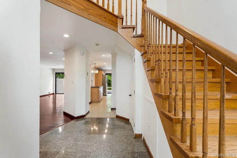 Entry foyer featuring speckled flooring, a natural wood staircase with balusters, recessed lighting, and a doorway opening to a room with dark wood-finish flooring Entry foyer featuring speckled flooring, a natural wood staircase with balusters, recessed lighting, and a doorway opening to a room with dark wood-finish flooring